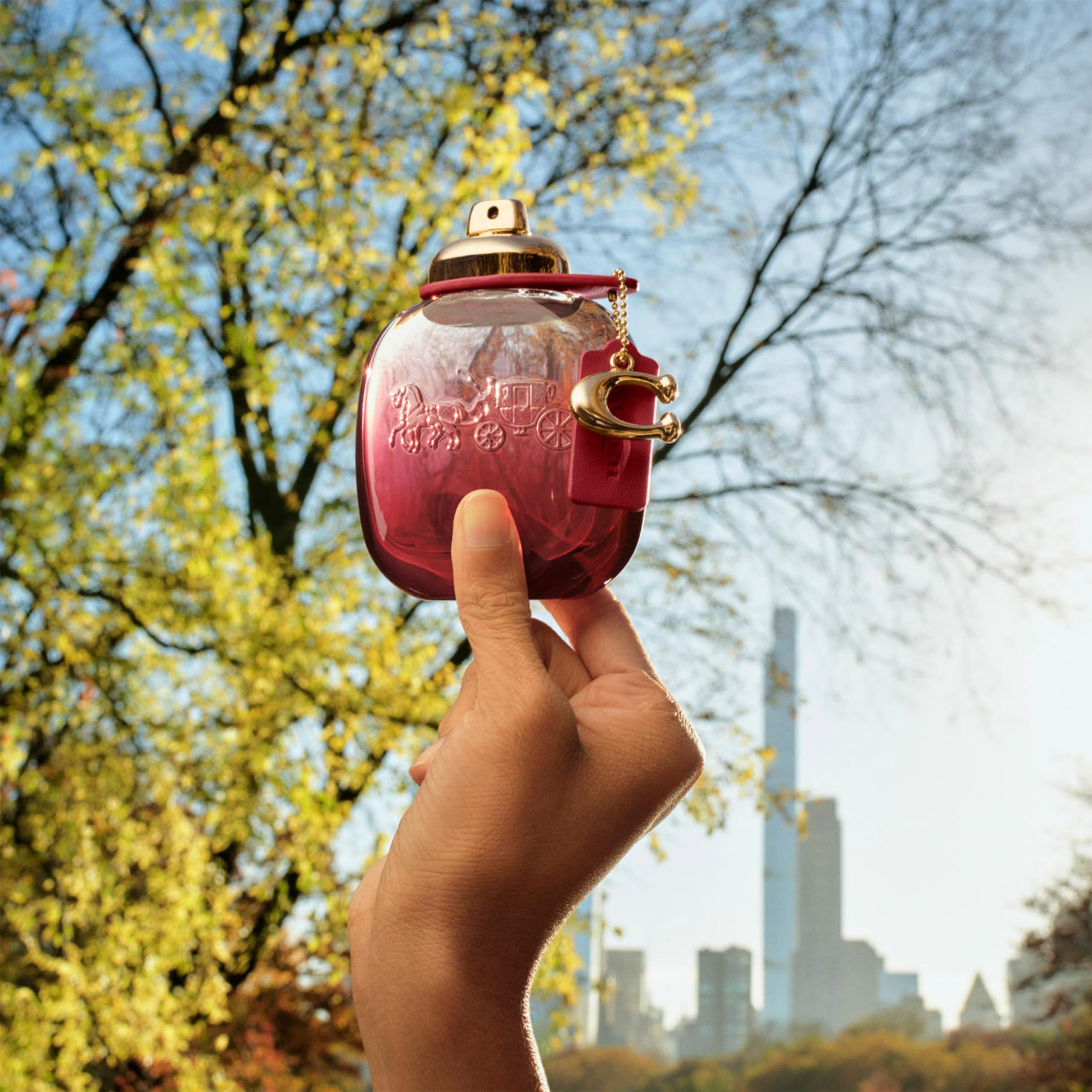 Una mano tiene un flacone davanti a uno sfondo di alberi ed edifici. Il flacone è rosa-rosso e ha un ciondolo dorato.