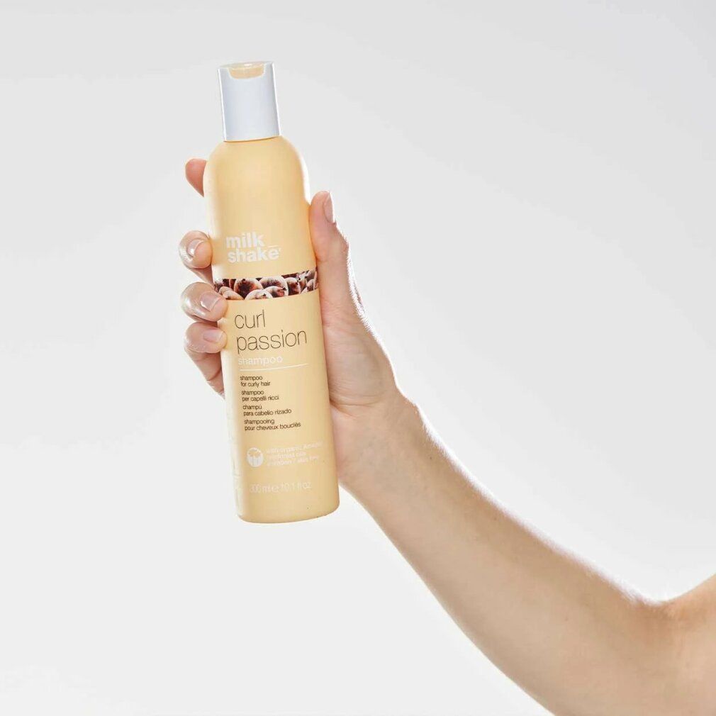 Mano che tiene un flacone di shampoo beige con tappo bianco. Scritta: milk_shake, curl passion shampoo. Per capelli ricci.