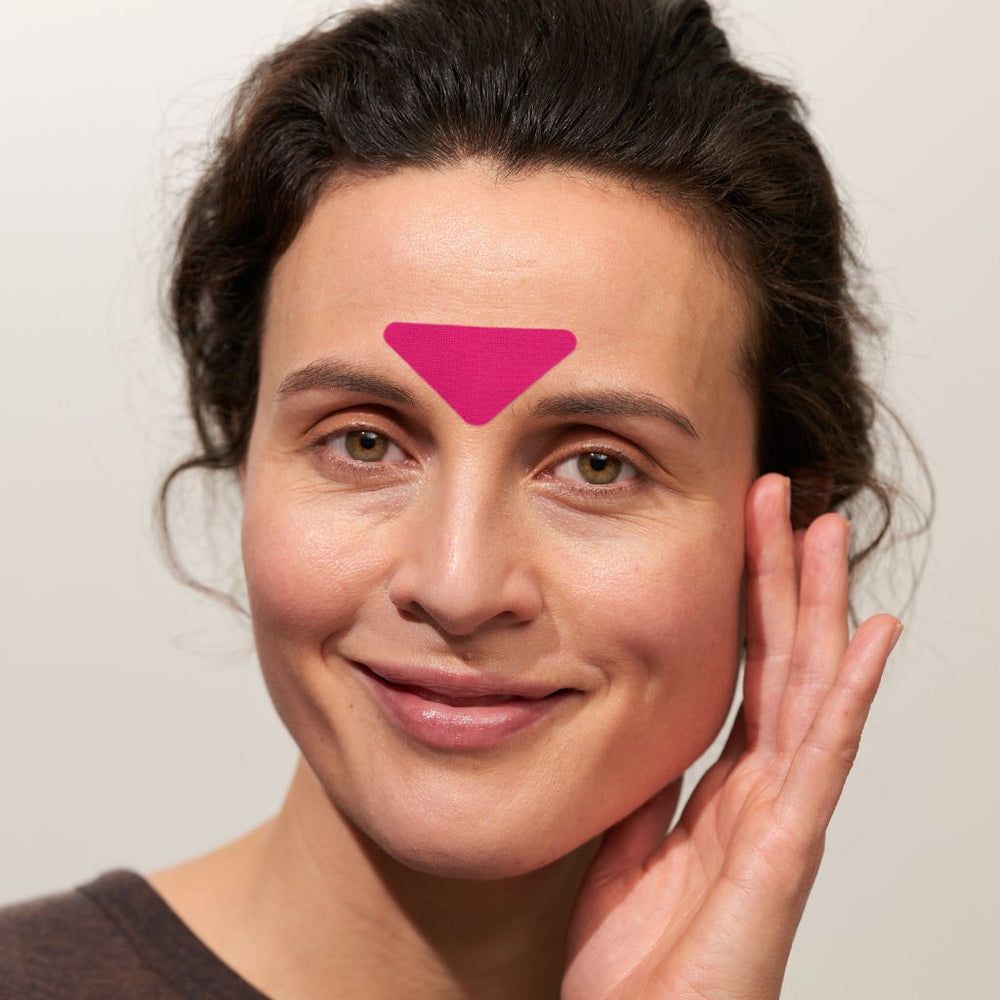 Primo piano di una donna con un cerotto rosa sulla fronte. Il cerotto è triangolare. La donna sorride.