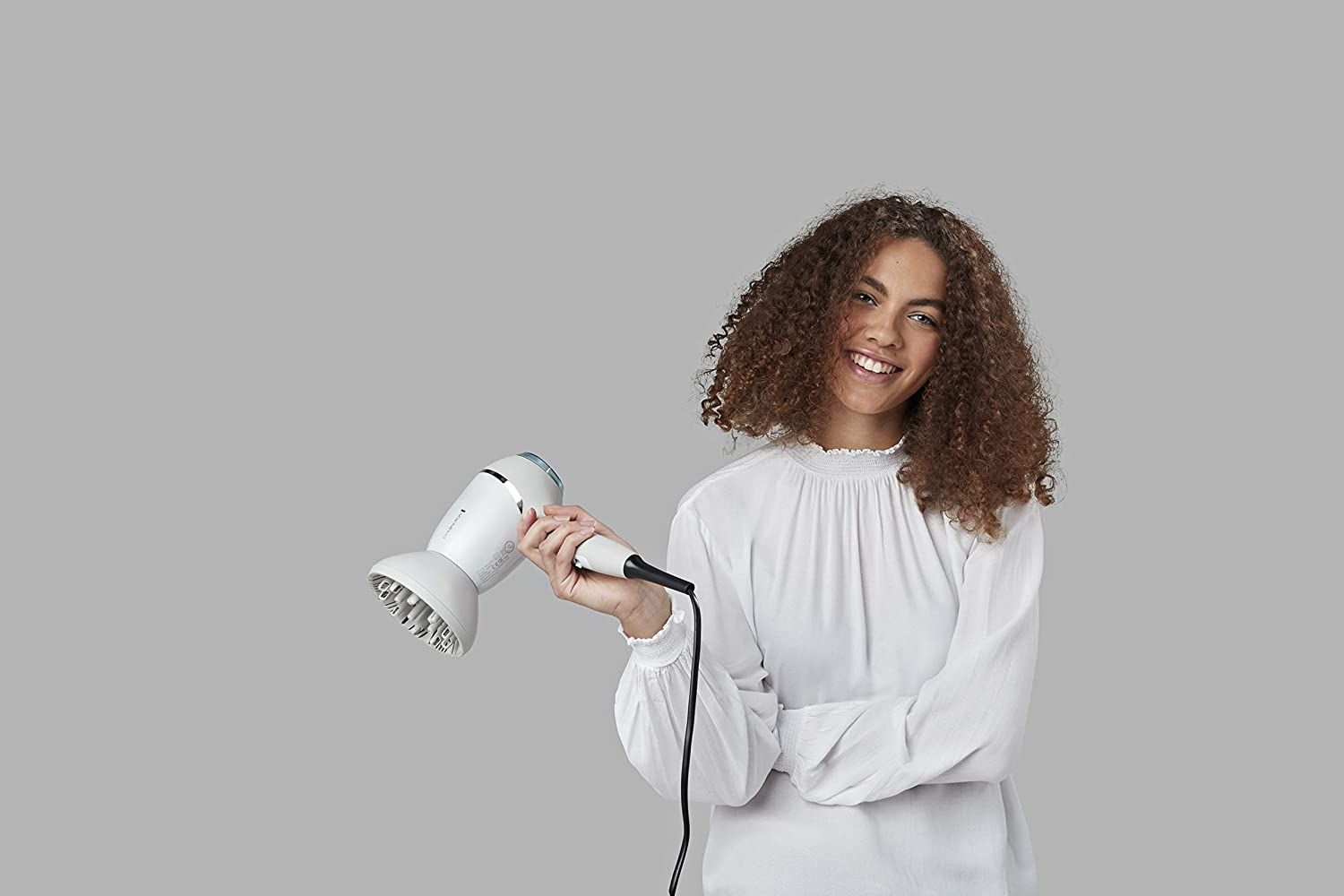 Donna tiene asciugacapelli bianco. Sfondo grigio. Indossa un top bianco.