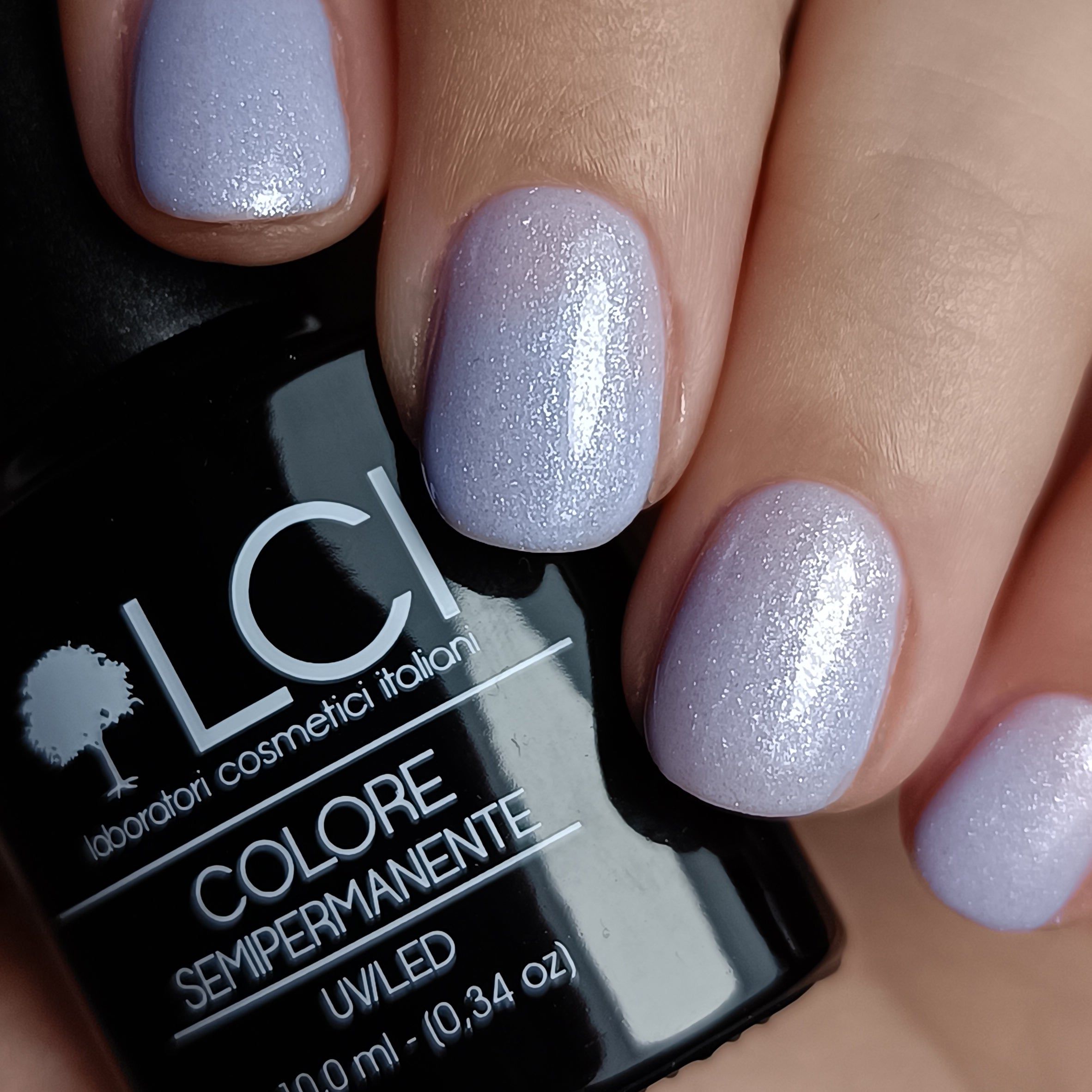 Mano con unghie smaltate e flacone di smalto. Unghie azzurro chiaro, glitterate. Flacone con testo, tappo nero.