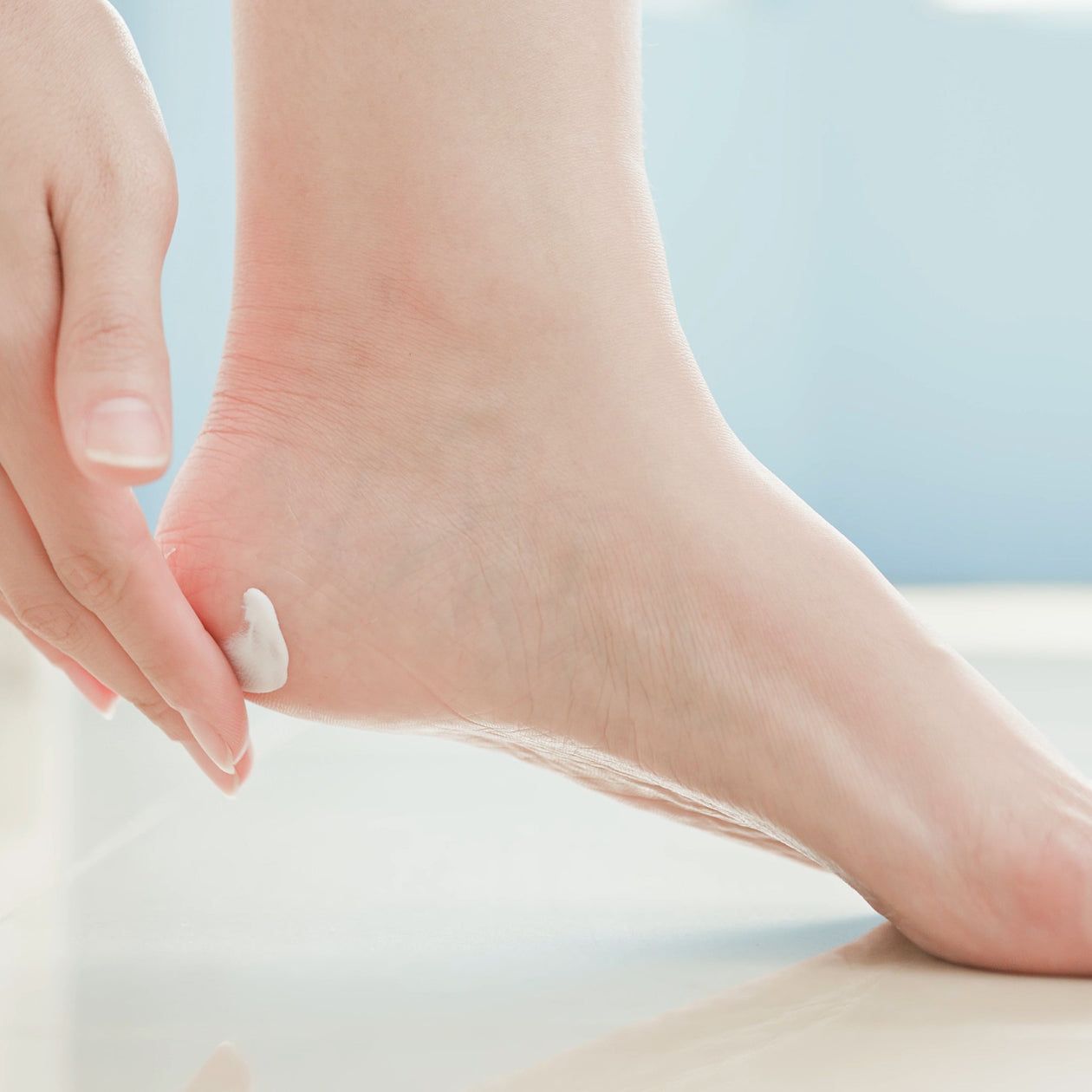 Primo piano di un piede. Una mano applica la crema sul tallone. Sfondo chiaro.