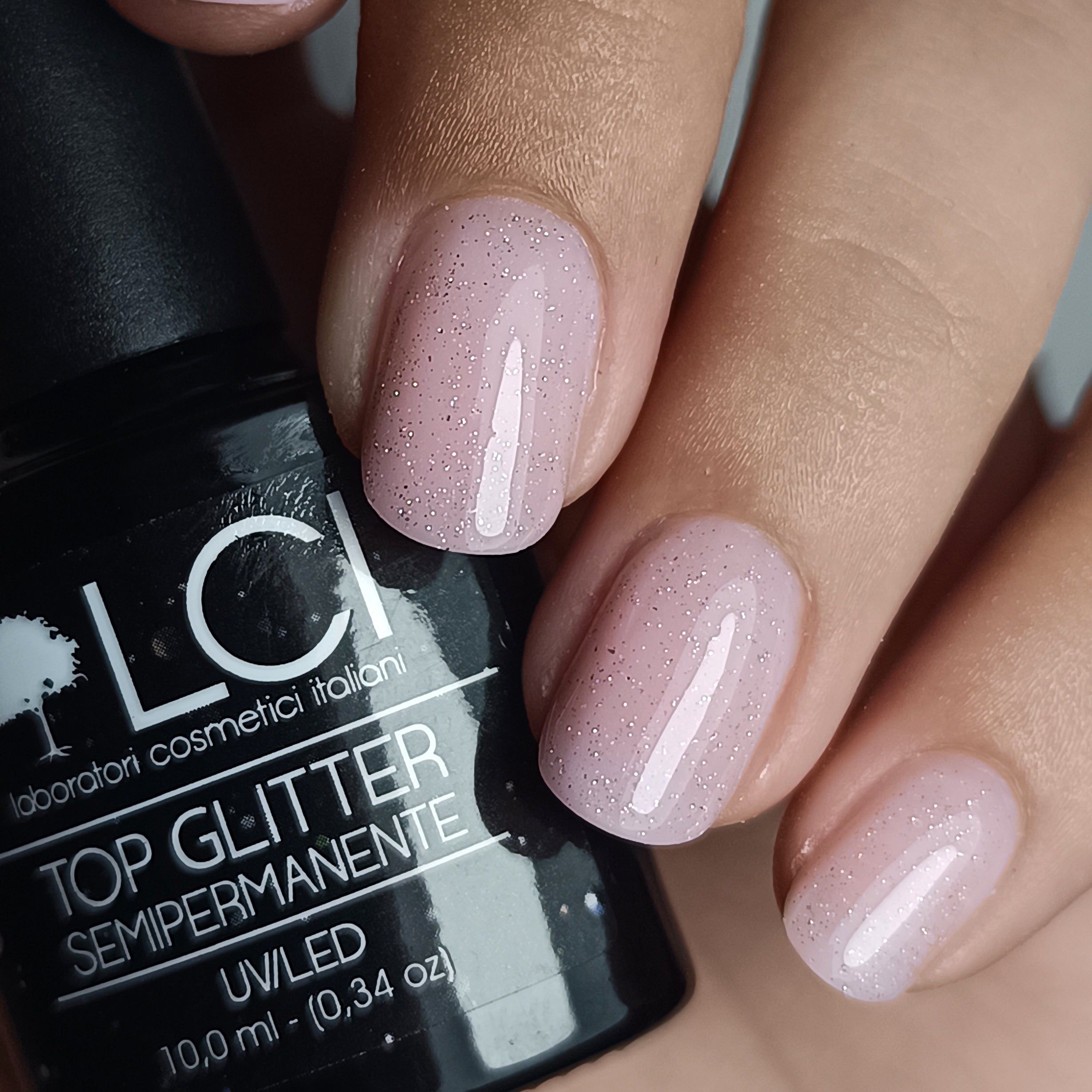 Mani con unghie smaltate. Top coat glitterato rosa. Flacone nero LCI Cosmetics sullo sfondo.