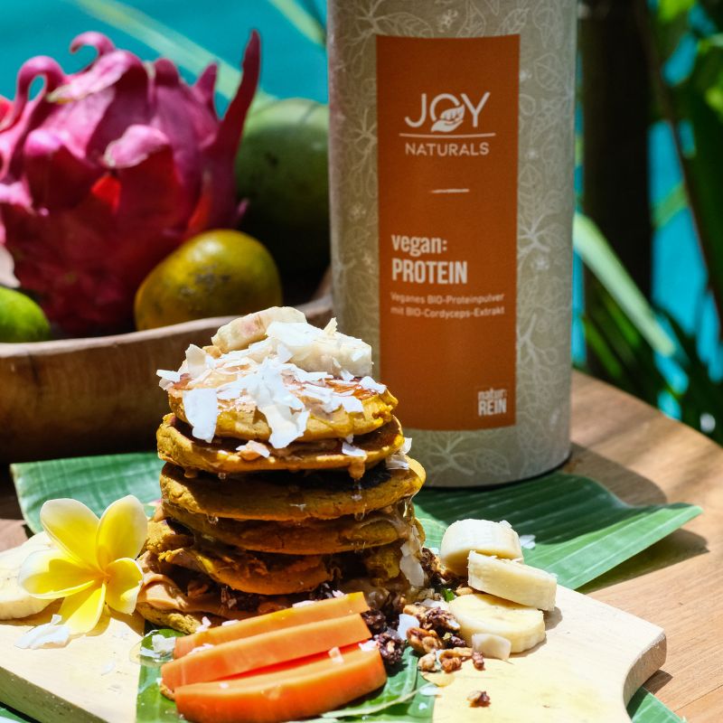 Barattolo accanto a pancake, frutta e fiori. Scritta: JOY NATURALS, vegan:PROTEIN. Complesso proteico BIO vegano con estratto di Cordyceps BIO.