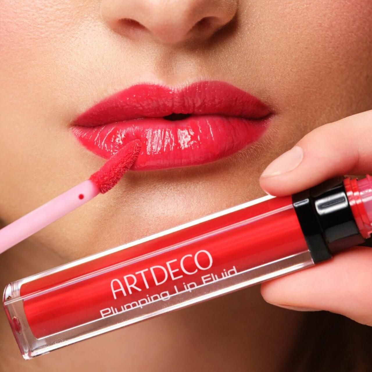 Applicazione di Artdeco Plumping Lip Fluid sulle labbra. Labbra rosse, prodotto e applicatore visibili. Nome del prodotto.