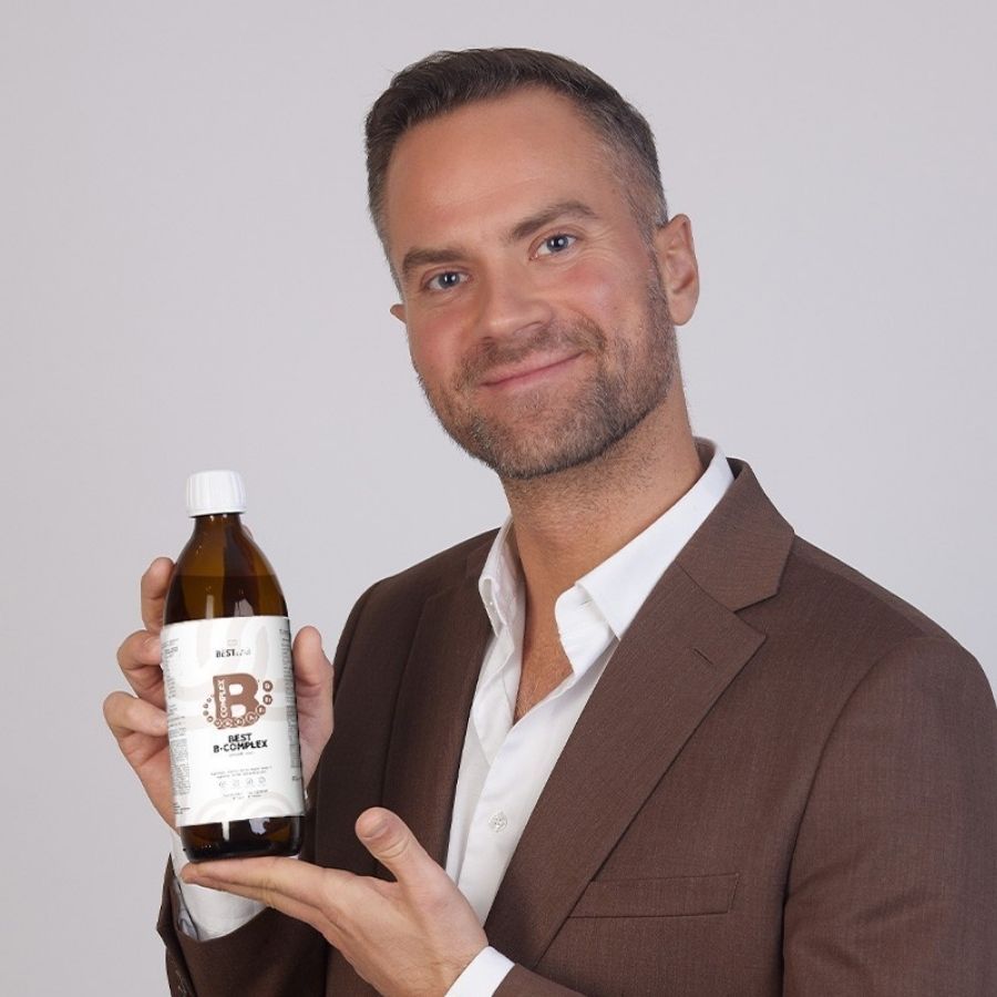 Uomo tiene bottiglia marrone "BEST LAB B-COMPLEX". Indossa un abito marrone. Sfondo chiaro.
