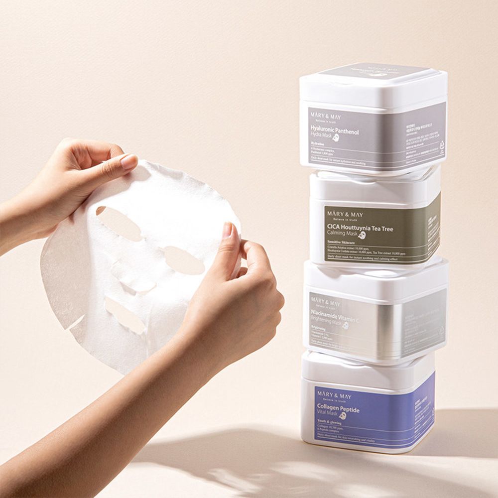 Mani che tengono una maschera per il viso. Pila di contenitori di maschere MARY & MAY: Hyaluronic Panthenol, CICA, Niacinamide, Collagen Peptide.