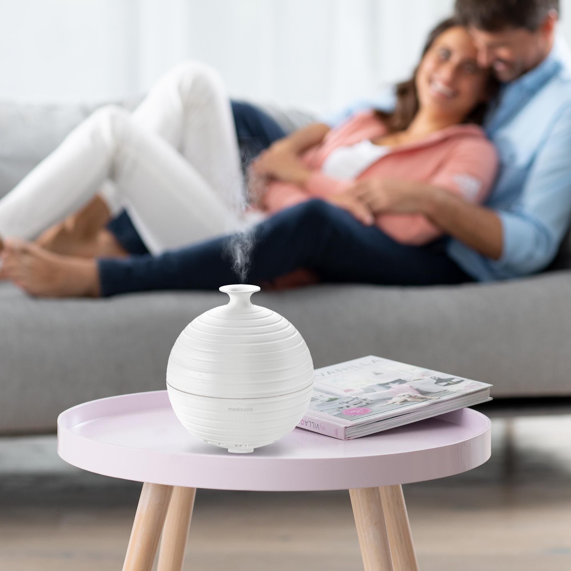 Diffusore di aromi su un tavolino, con una coppia che si rilassa su un divano sullo sfondo.