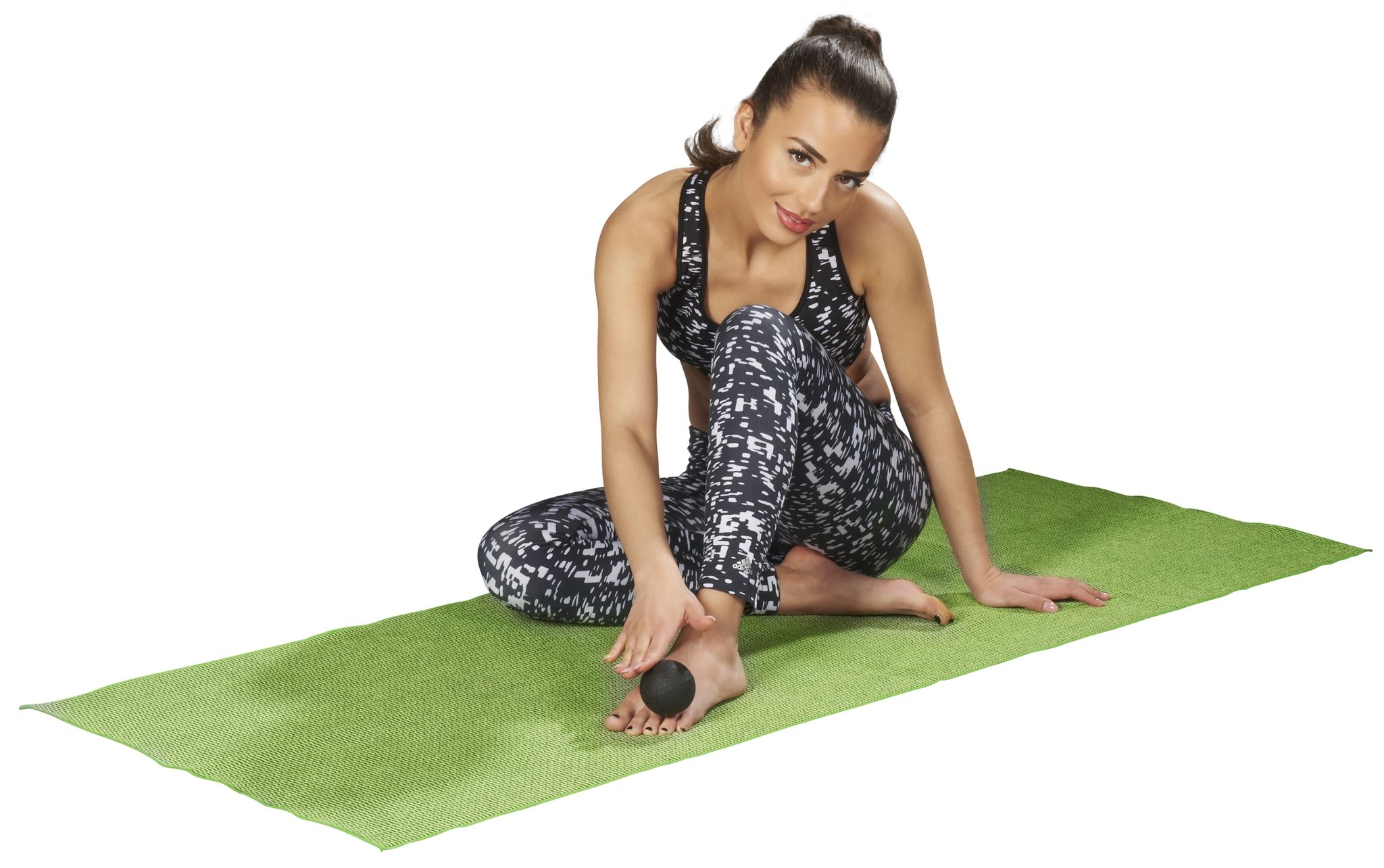Donna seduta su un tappetino verde. Pallina nera sotto il piede. Abbigliamento sportivo: top e pantaloni fantasia. Guarda la fotocamera.