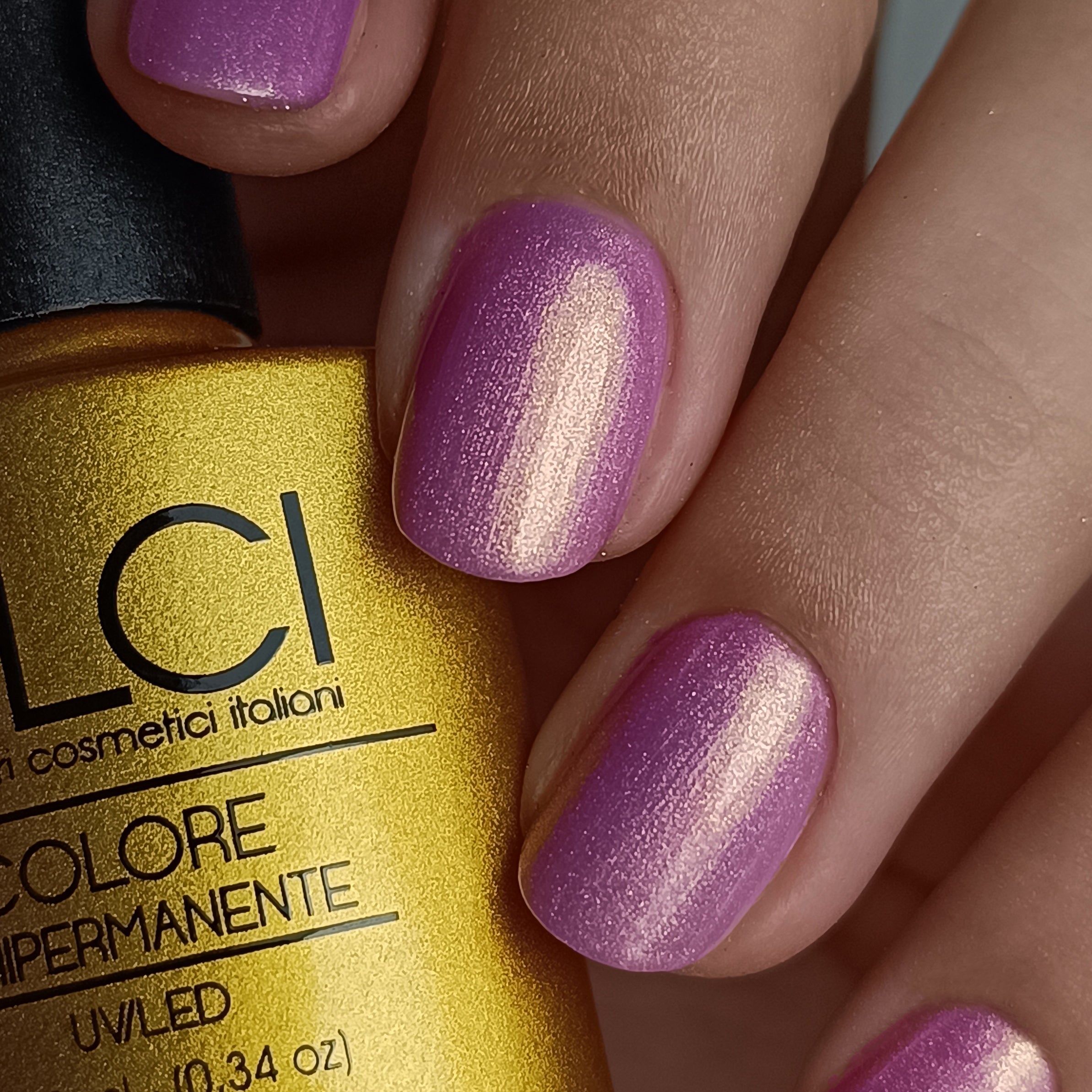 Mano con unghie laccate rosa-oro. Flacone con etichetta LCI sullo sfondo.