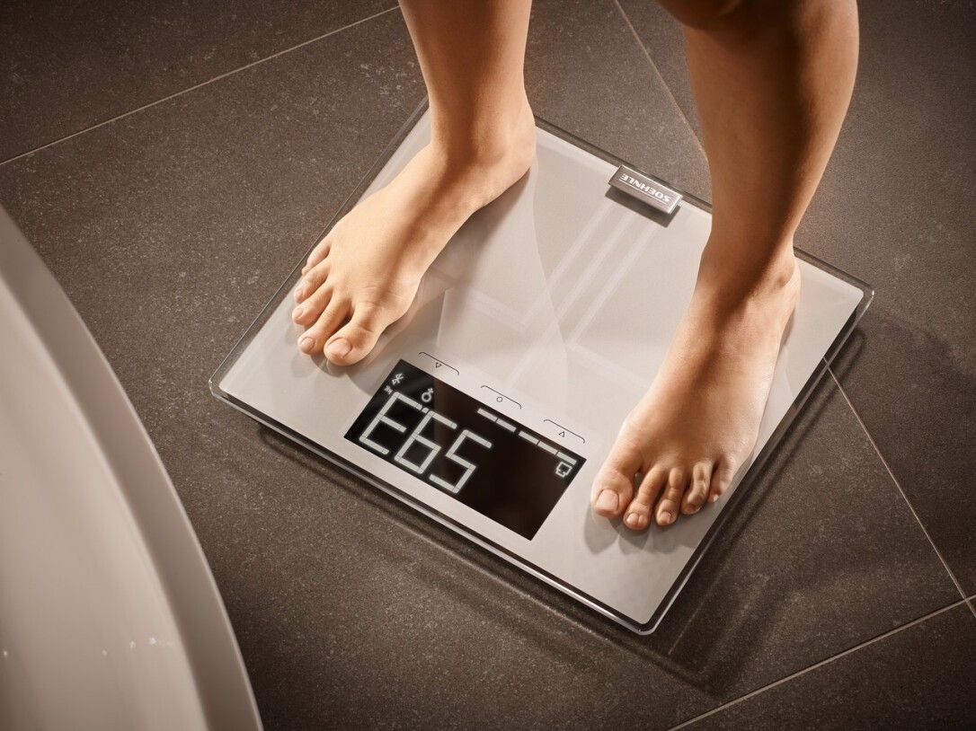 Piedi su bilancia. Bilancia grigia con display digitale. Mostra il peso. Su pavimento piastrellato.
