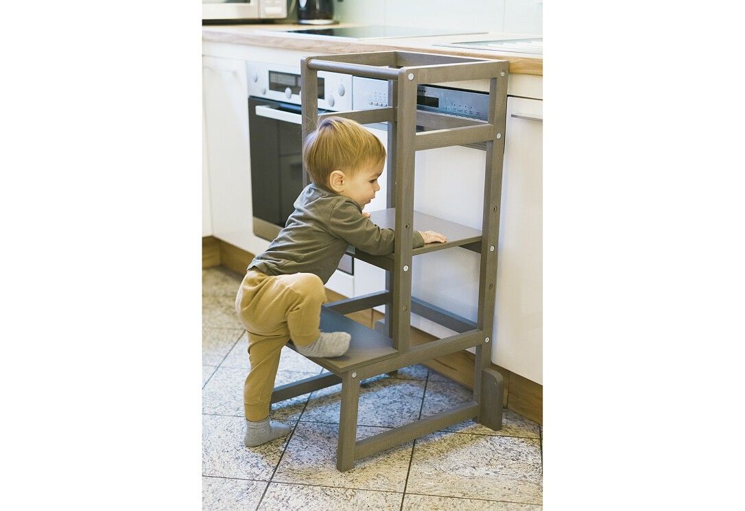 Bambino che sale su una torre di apprendimento grigia in cucina. Il bambino si sostiene.