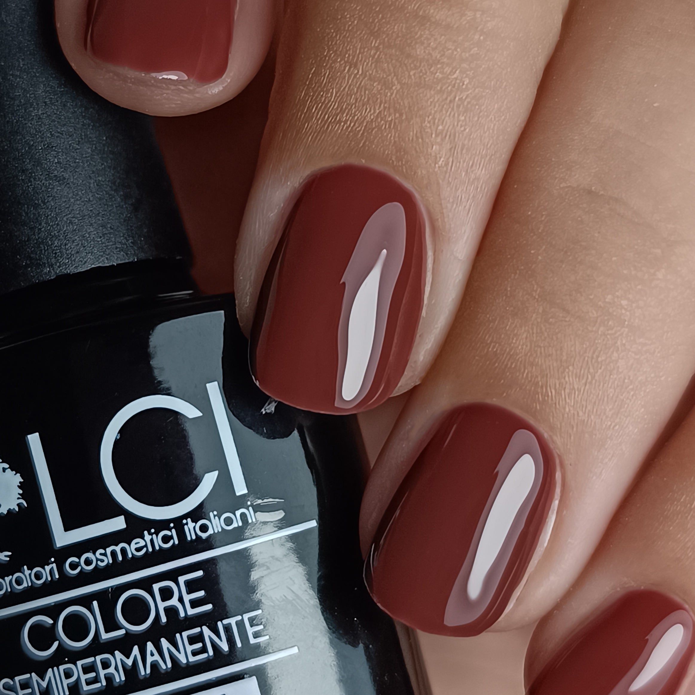 Primo piano di unghie smaltate di marrone. Flacone parzialmente visibile. Unghie di forma ovale, lucide.