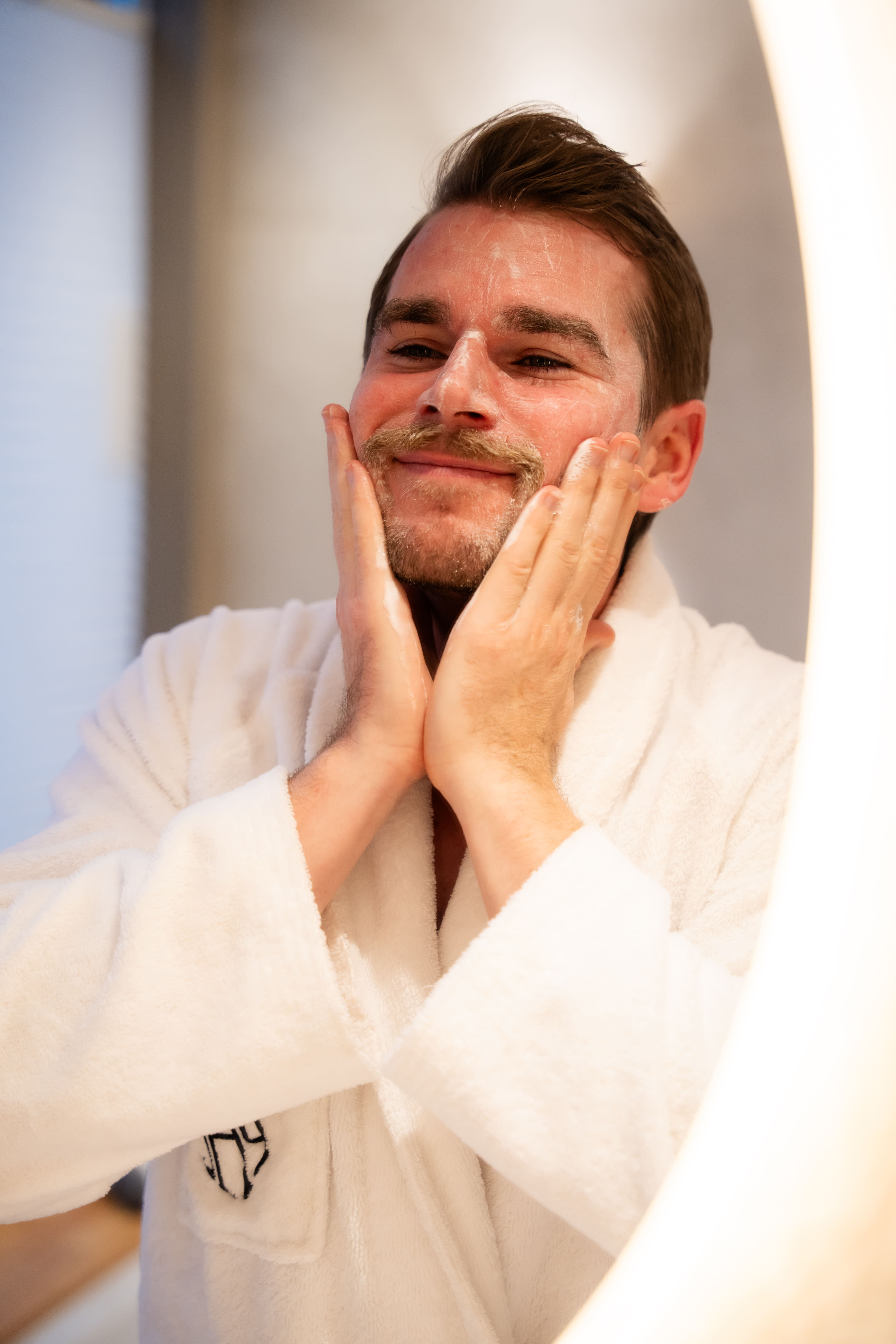 Uomo in vestaglia bianca si massaggia il viso davanti a uno specchio illuminato.