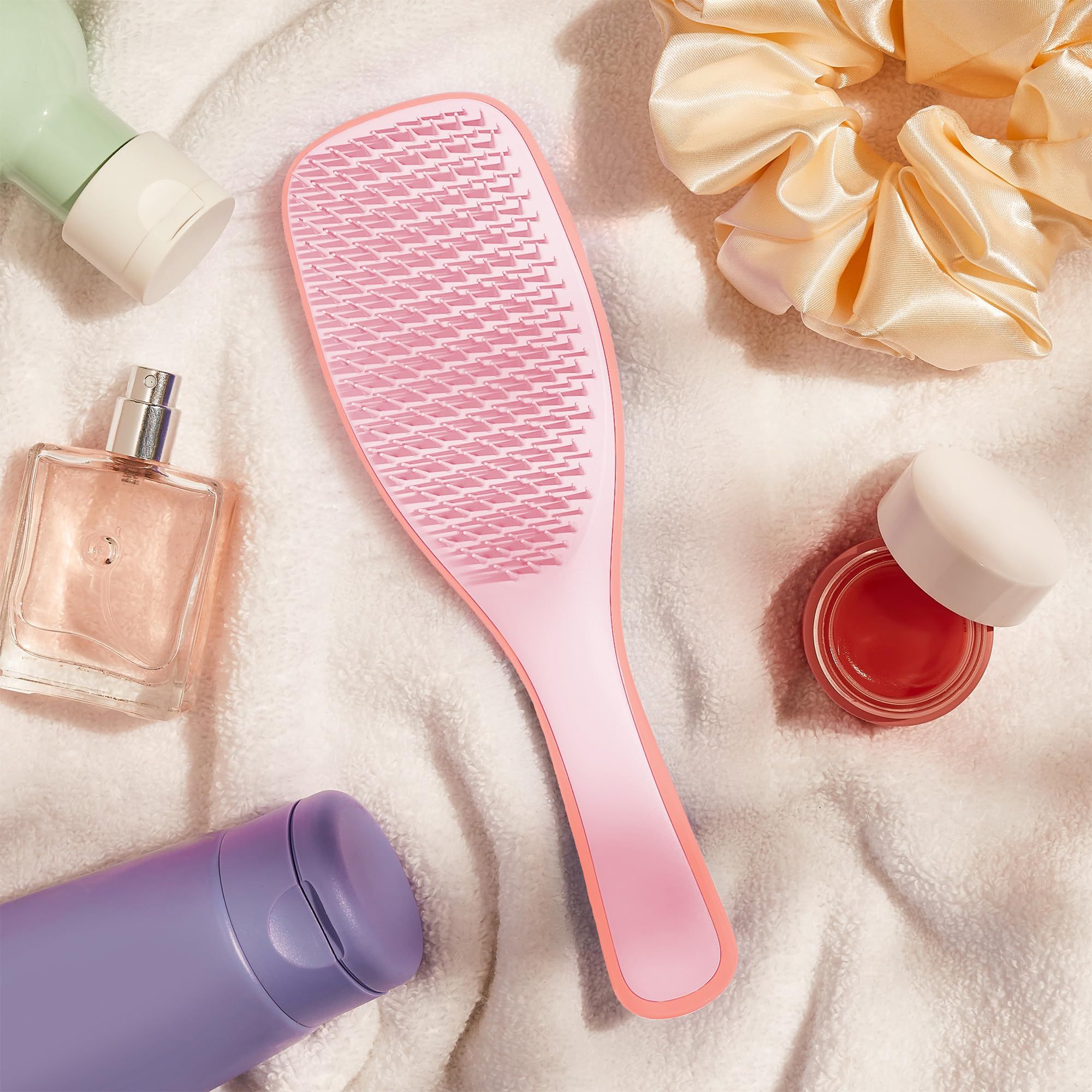 Spazzola rosa su un asciugamano bianco, circondata da prodotti cosmetici: profumo, balsamo per le labbra, olio per capelli e lacca.