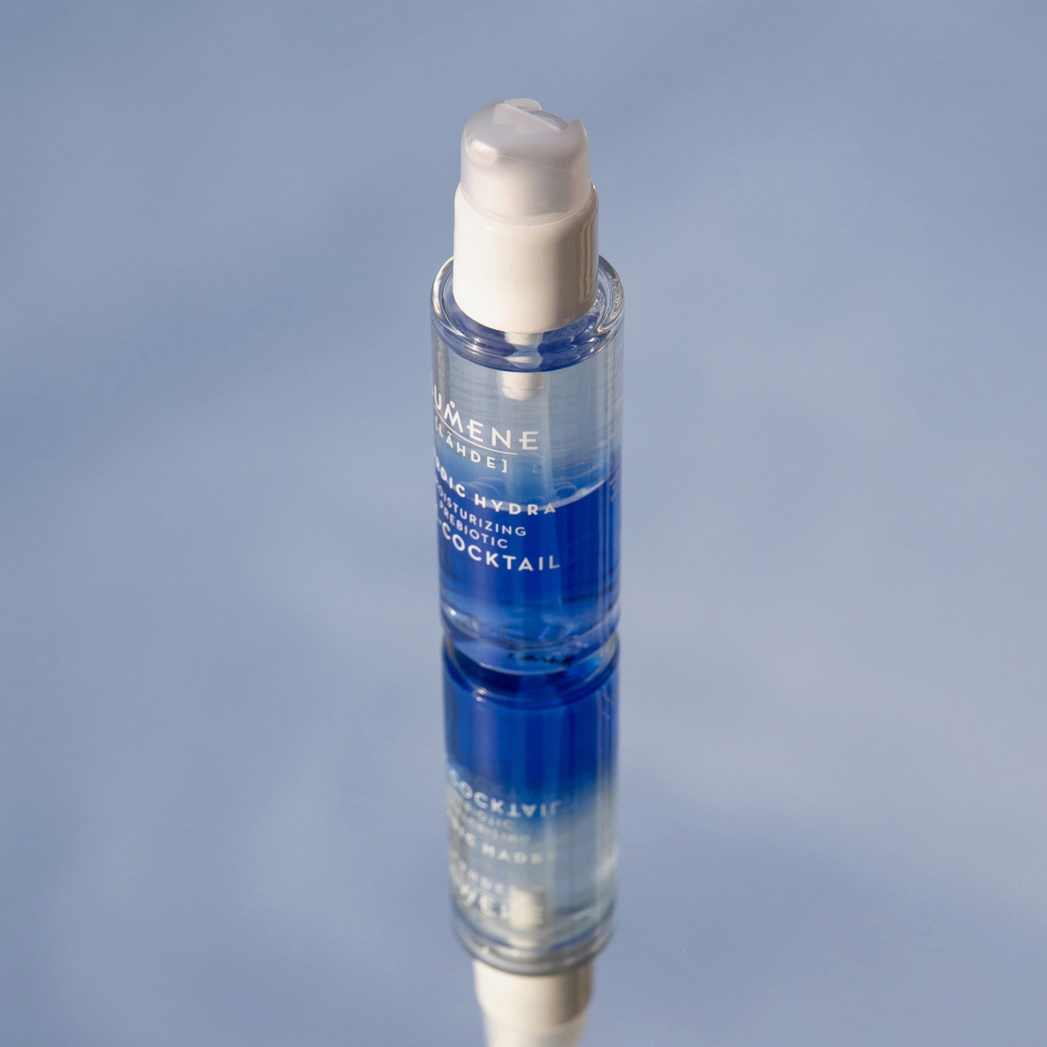Flacone trasparente con erogatore bianco, su superficie riflettente. Liquido blu e trasparente visibile. Nome del prodotto e marchio.