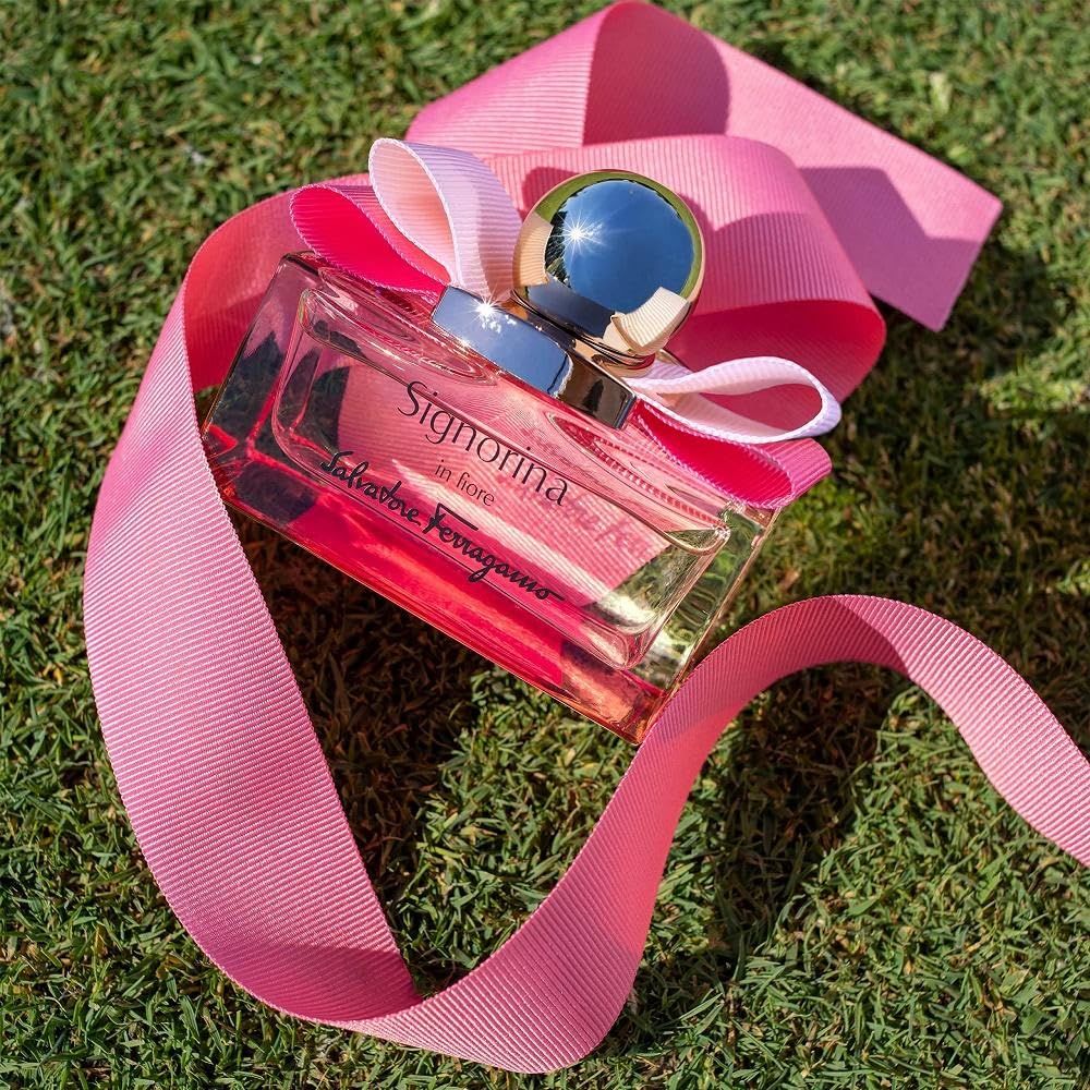Flacone in vetro con liquido rosa, fiocco rosa e tappo dorato, su un nastro rosa ed erba verde. Scritta Signorina in fiore.