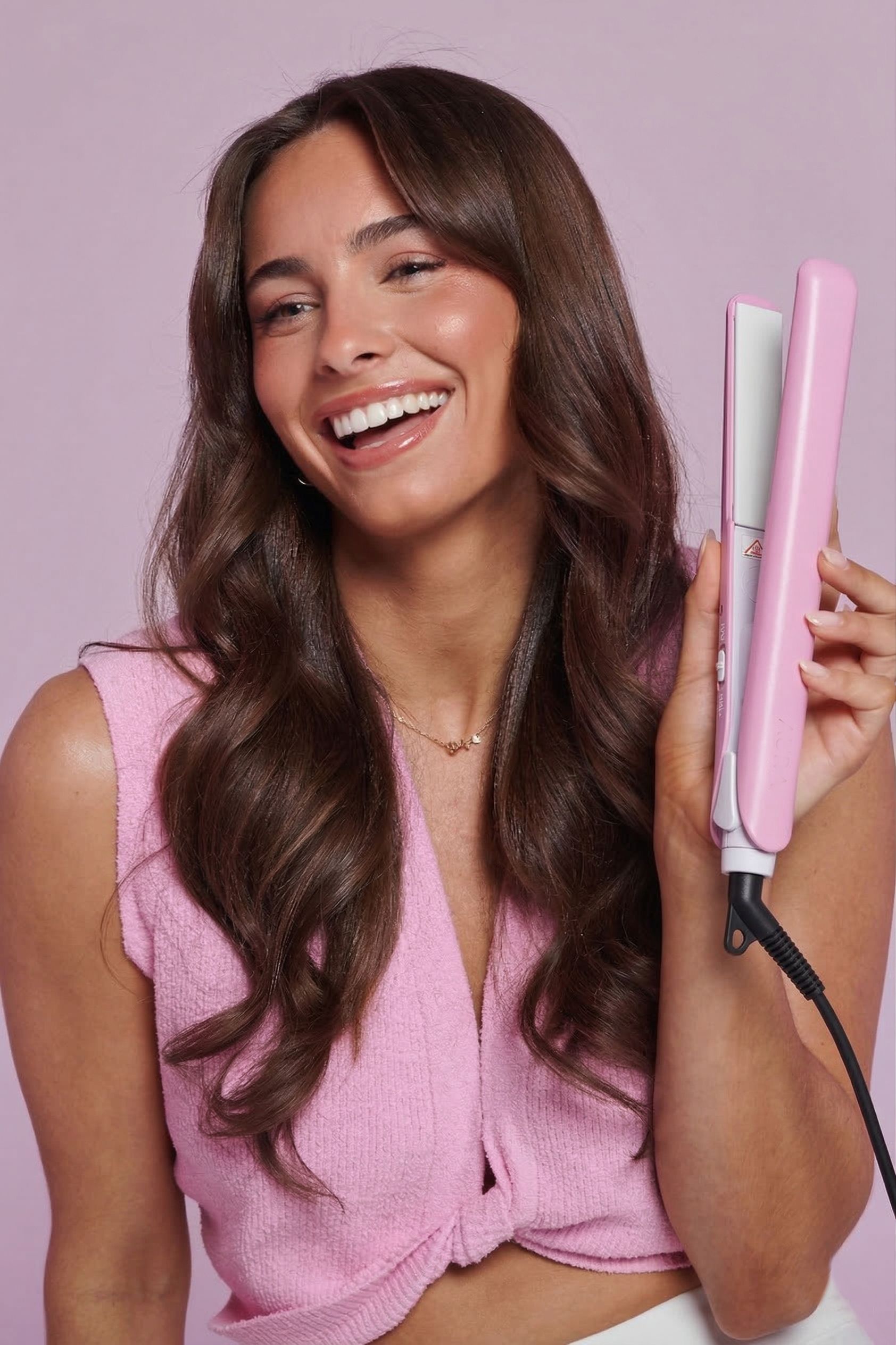 Donna tiene piastra rosa. Cavo nero. La donna sorride. Capelli castani mossi. Top rosa.