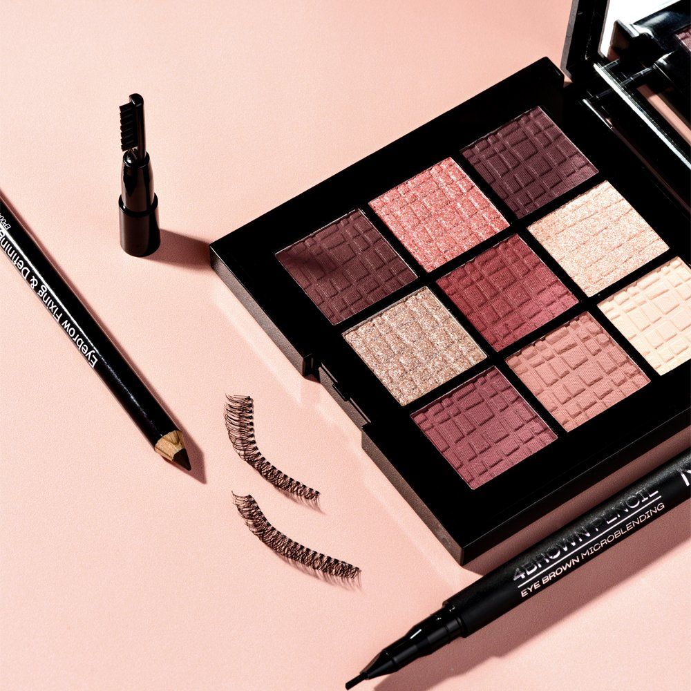Palette ombretti aperta con 9 colori, accanto ciglia finte, matita per sopracciglia e eyeliner. Sfondo rosa.