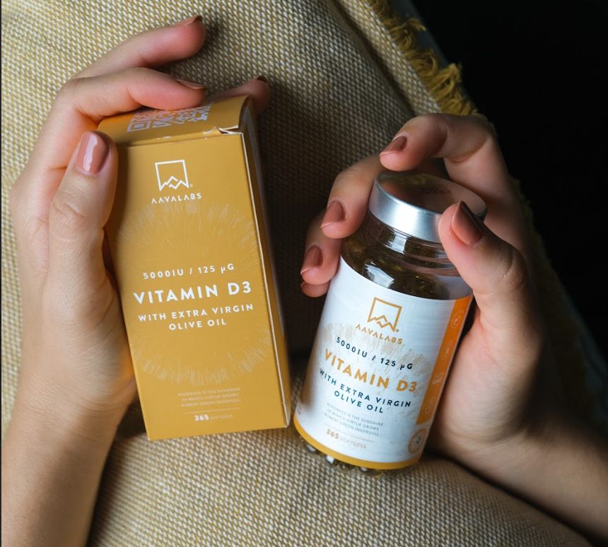 Mani che tengono una scatola e un flacone. Scritta: Vitamina D3, 5000 UI, con olio d'oliva extra vergine. Sfondo giallo.