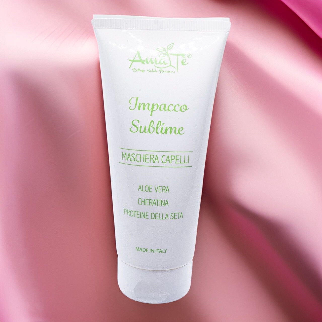 Tubo bianco con testo verde: Amatè, Impacco Sublime, Maschera Capelli. Aloe Vera, Cheratina, Proteine della seta. Su tessuto rosa.