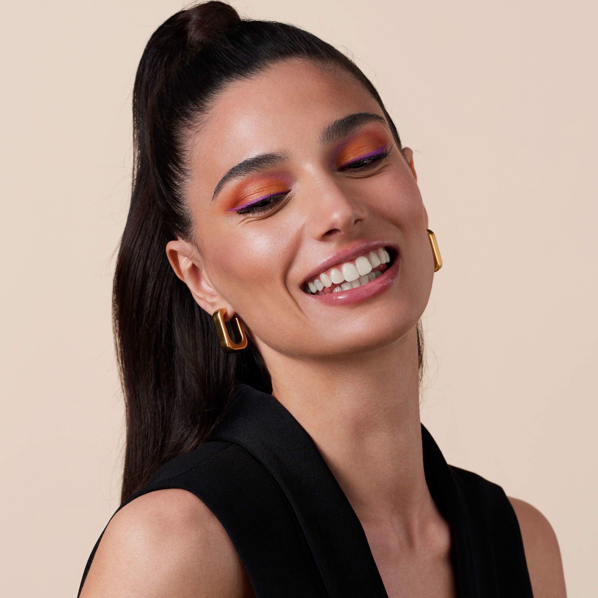 Una donna sorridente con i capelli raccolti e orecchini d'oro. Indossa un top nero e trucco.