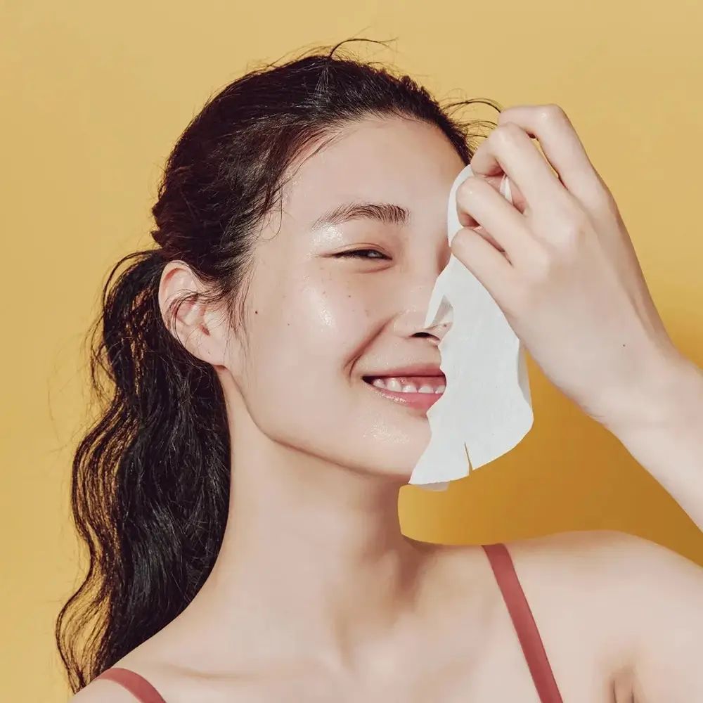 Persona che tiene una maschera per il viso davanti al viso. La maschera copre metà del viso. Sfondo giallo. La persona sorride.