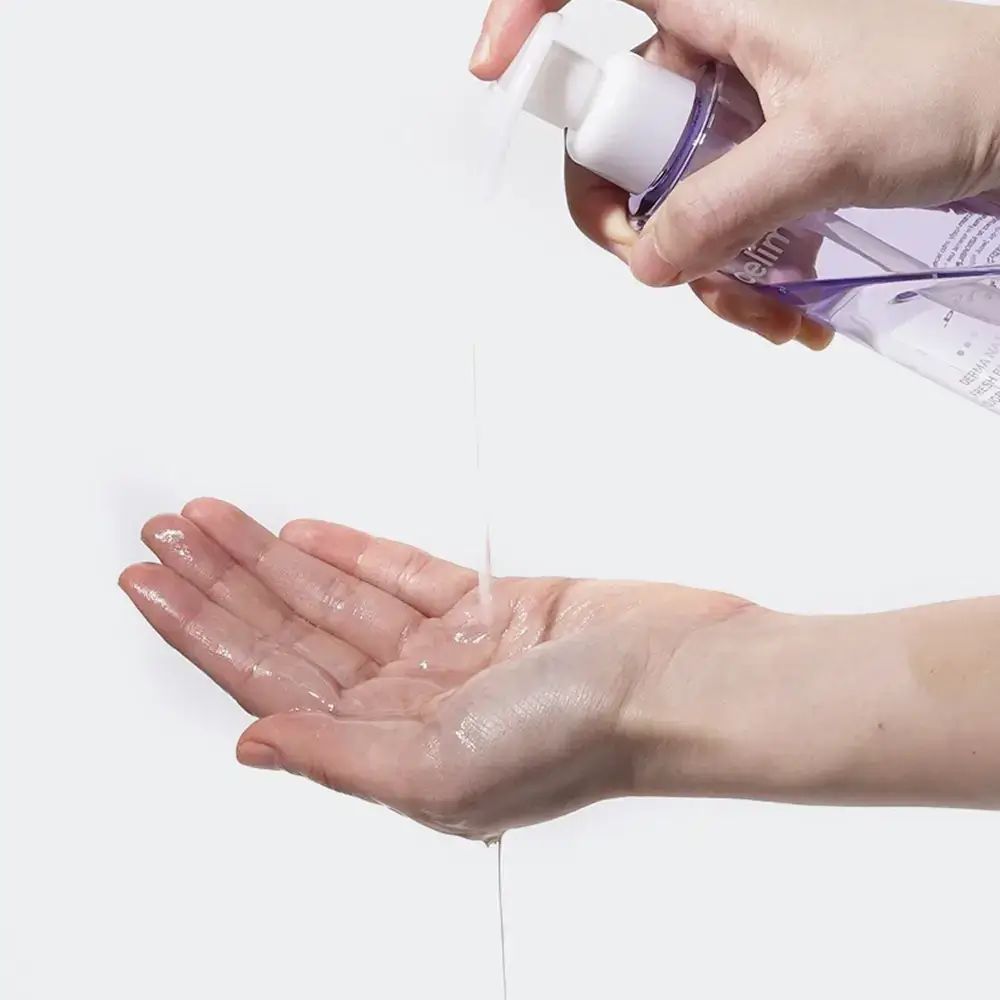 Mani, una tiene una bottiglia, l'altra riceve liquido. La bottiglia ha un erogatore bianco e dettagli viola. Il liquido scorre nella mano.