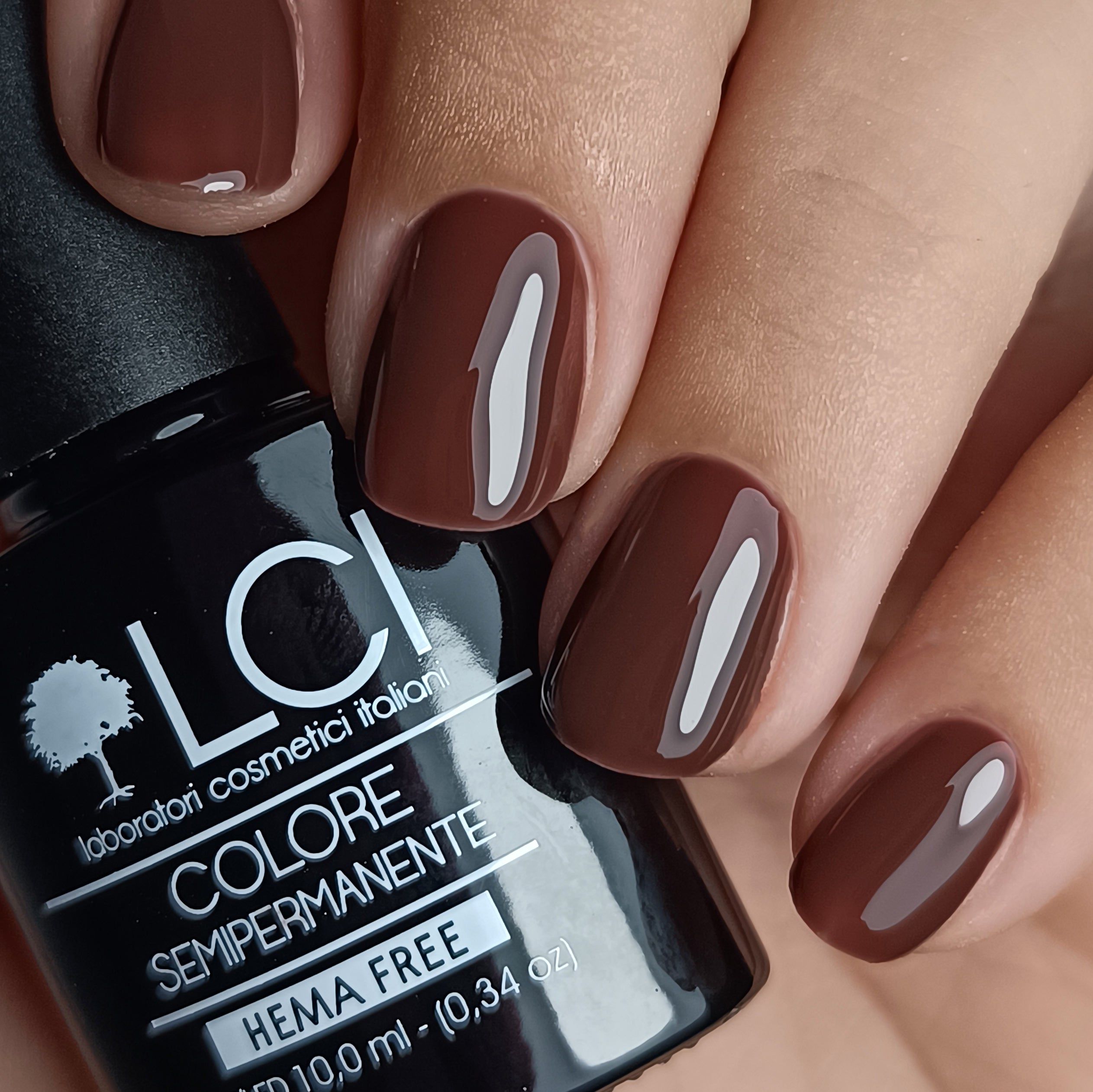 Mano con unghie smaltate marrone scuro. Flacone di smalto accanto. Scritta: LCI, Colore Semipermanente.