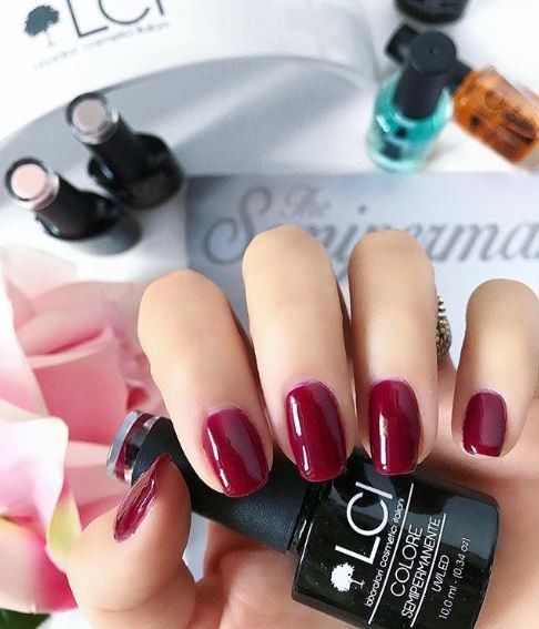 Mano con unghie smaltate e flacone di smalto. Smalto rosso scuro. Scritta: LCI, Colore Semipermanente, UVLED 10,0 ml.