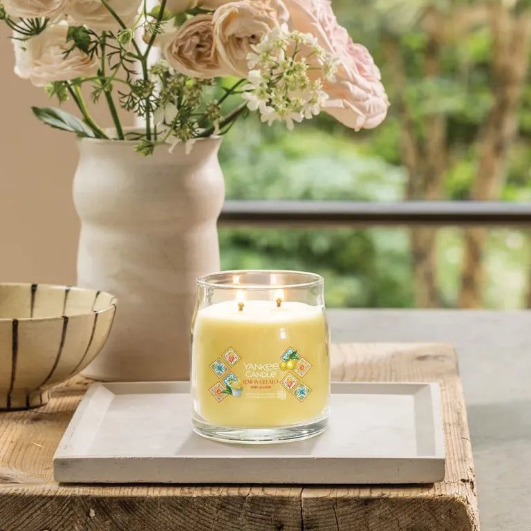 Candela in vetro con cera gialla, due stoppini ed etichette decorative. Su un vassoio, circondata da fiori e un vaso.