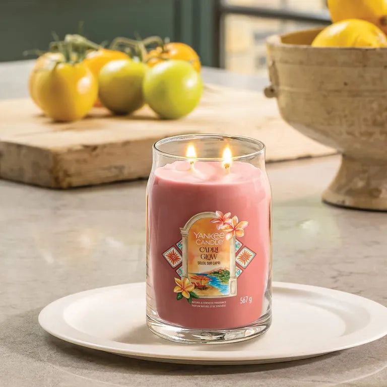 Candela rosa in vetro, due stoppini, su piatto bianco. Scritta: Capri Glow. Sfondo: pomodori.