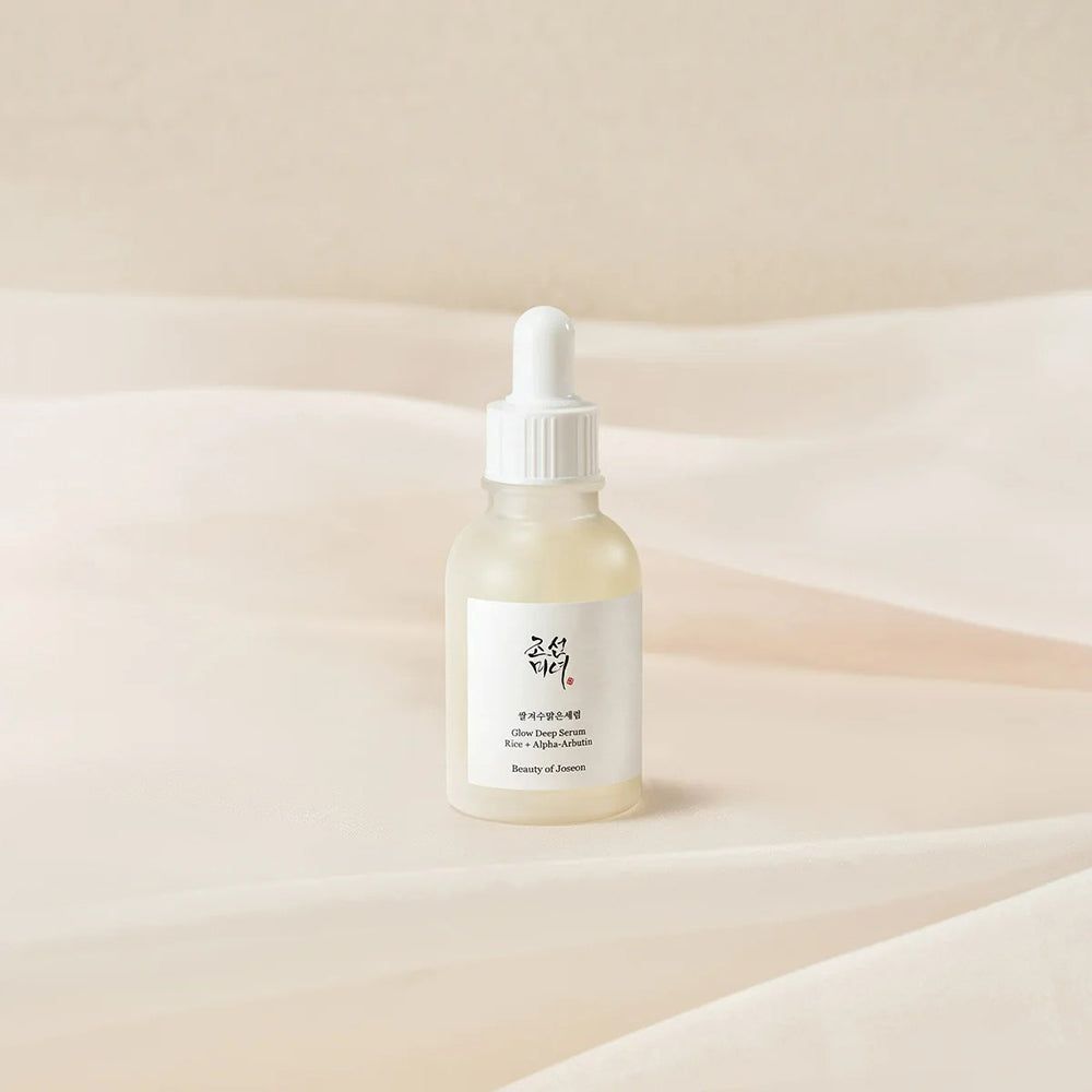 Flacone di siero con etichetta bianca. Scritta: Glow Deep Serum, Rice + Alpha-Arbutin. Marchio: Beauty of Joseon. Su tessuto chiaro.