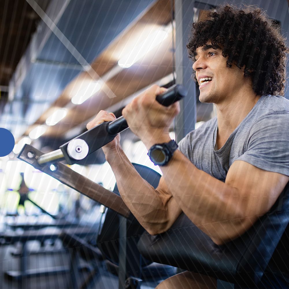 Uomo che si allena in palestra. Usa una macchina. Sorride. Indossa una maglietta grigia e uno smartwatch.