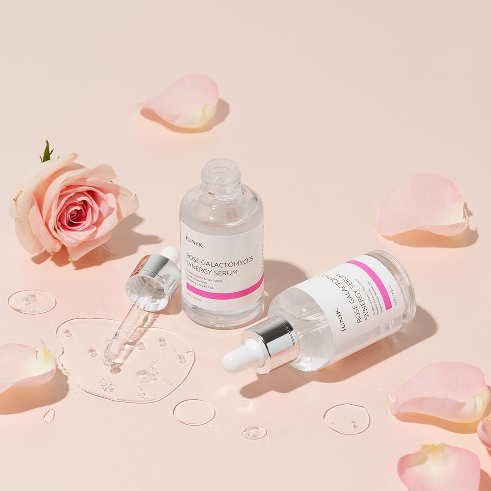 Due flaconi con pipette, petali di rosa e una rosa. Scritta: ROSE GALACTOMYCES SYNERGY SERUM. Liquido sulla superficie.