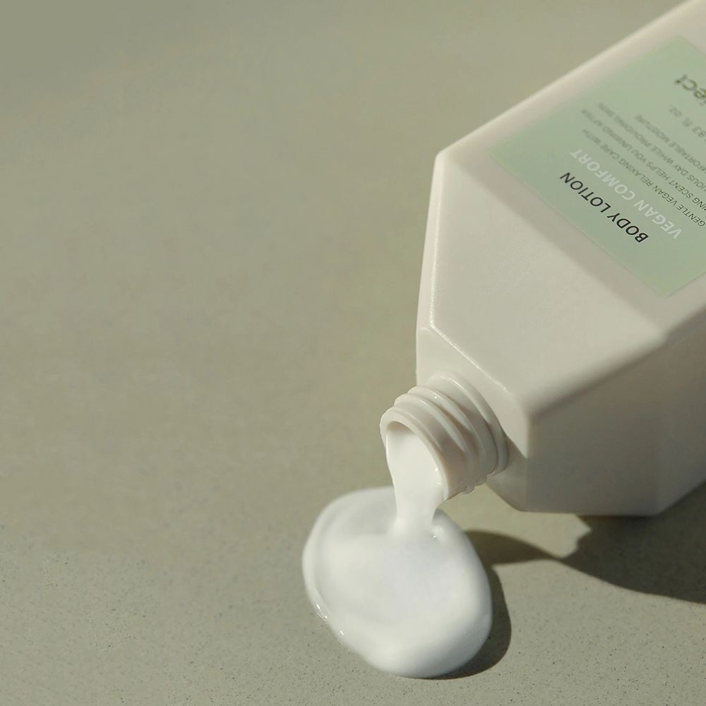 Lozione per il corpo versata da un flacone bianco. Scritta: Vegan Comfort, b:and project. Prodotto bianco su superficie grigia.
