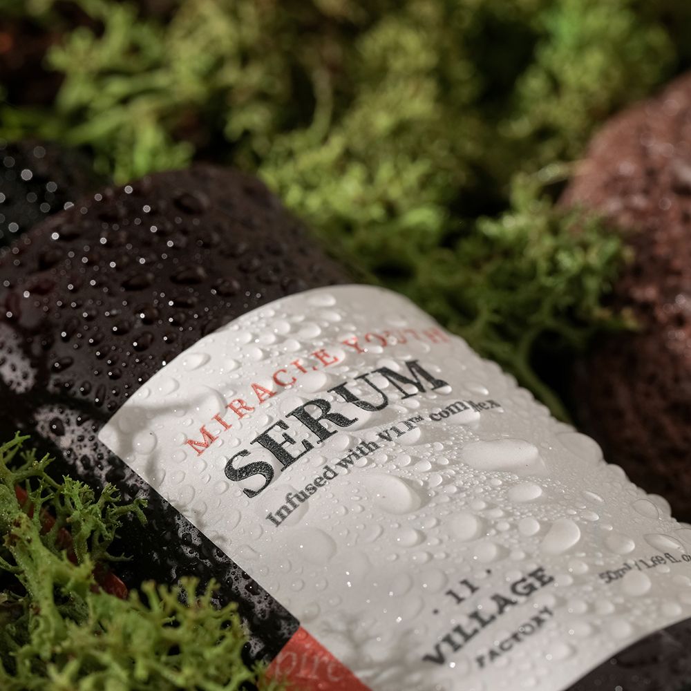 Flacone di siero marrone con etichetta bianca, coperto da gocce d'acqua. Testo: MIRACLE YOUTH SERUM. VILLAGE 11 FACTORY. Su muschio.