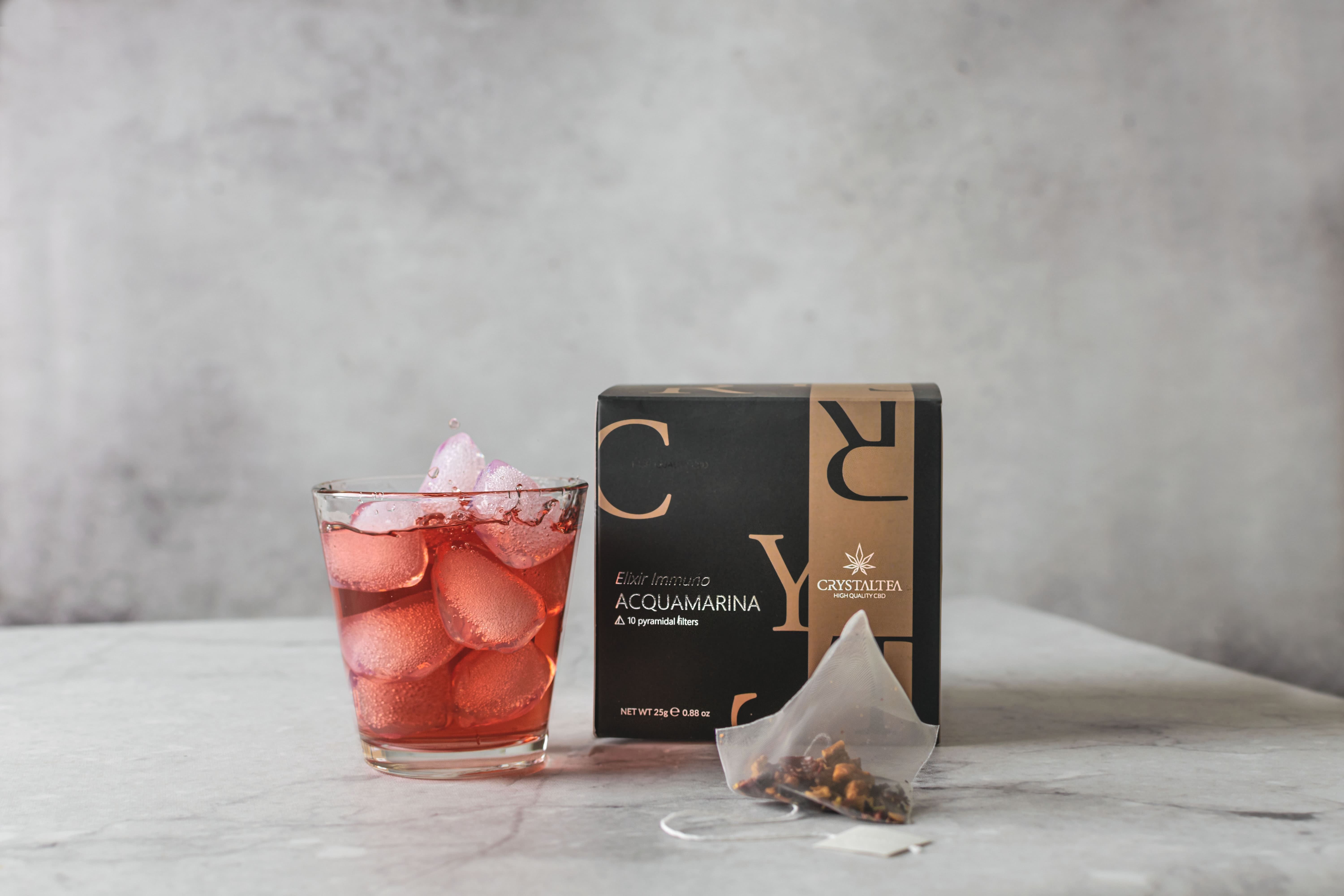 Bicchiere con cubetti di ghiaccio e bevanda rossa. Bustina di tè e scatola con logo. Sfondo grigio.