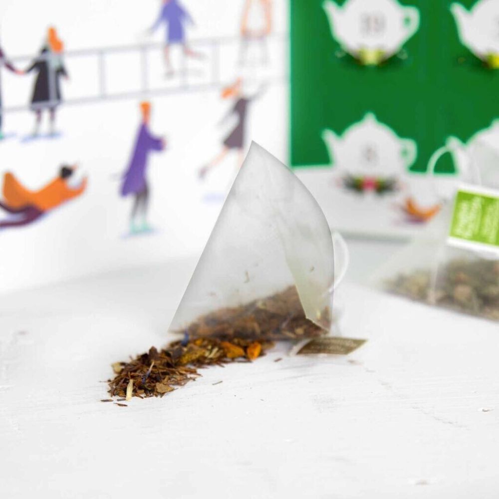 Bustina di tè aperta con foglie di tè sfuse. Sfondo: illustrazione del calendario dell'Avvento con teiere.