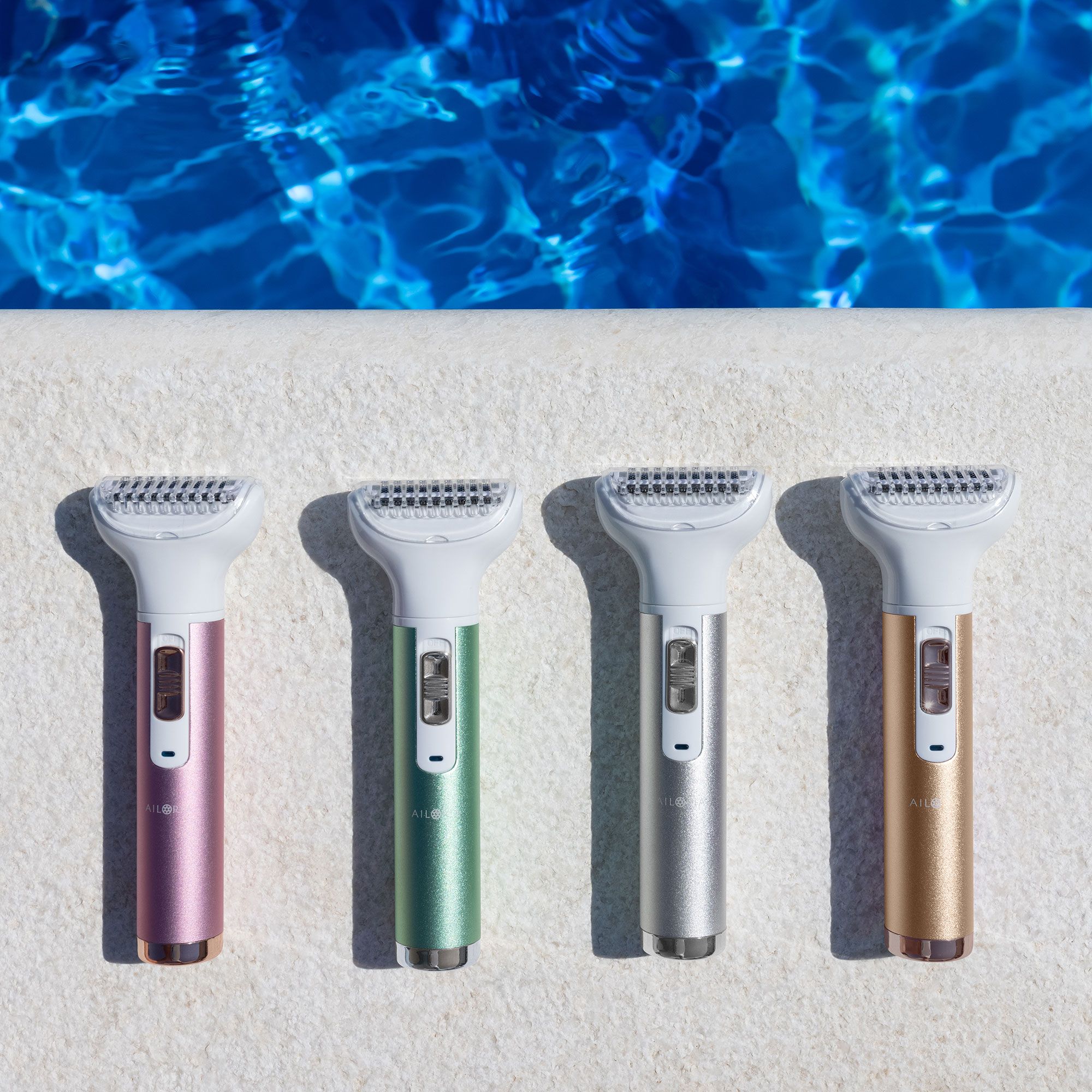 Quattro rasoi in diversi colori: rosa, verde, argento e oro. Posati su una superficie bianca vicino all'acqua blu.