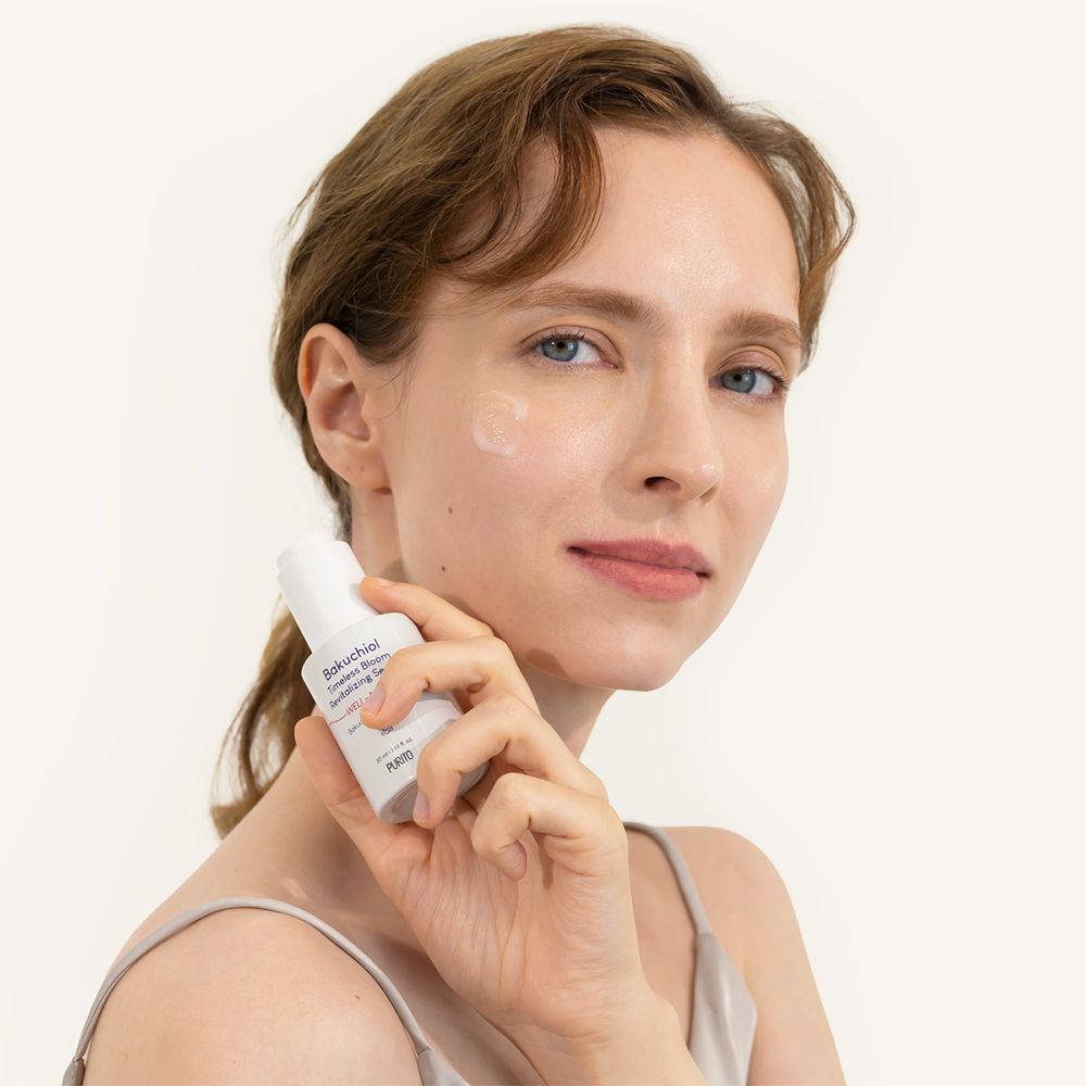 Donna che tiene in mano un flacone di siero bianco. Scritta: Bakuchiol Timeless Bloom Revitalizing Serum. Viso con siero.