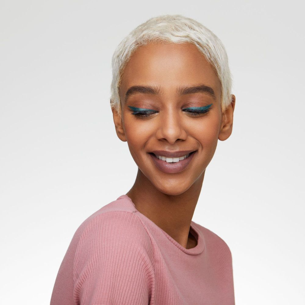 Donna con capelli biondi e eyeliner kajal. Indossa un top rosa e sorride.