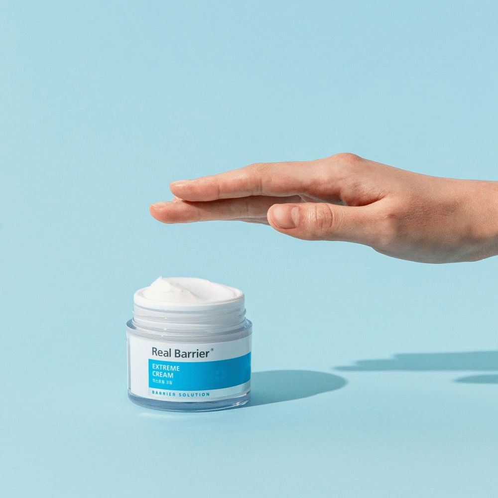 Mano sopra il vasetto di crema. Scritta: Real Barrier, Extreme Cream. Colori blu e bianco. Pelle chiara.