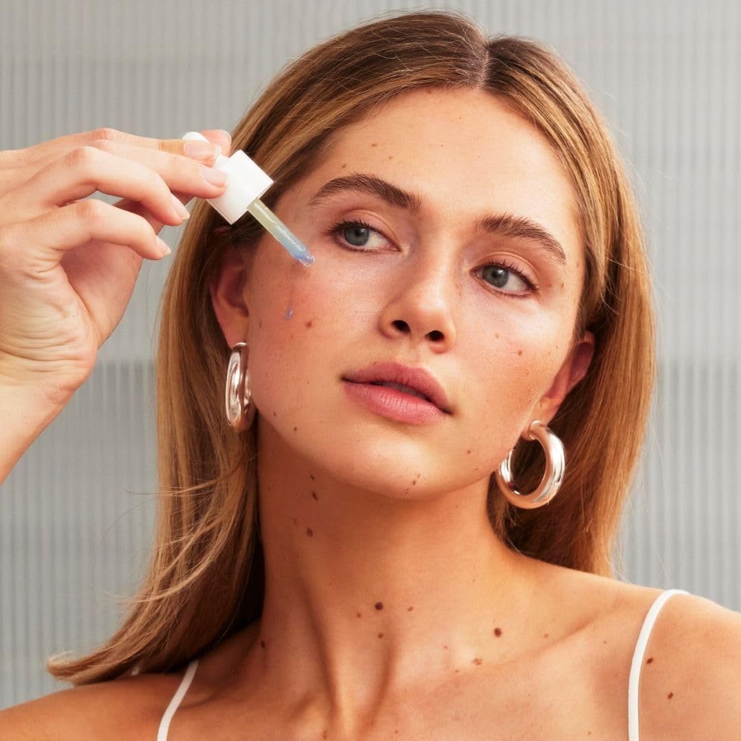 Donna con orecchini applica un prodotto sul viso con una pipetta. Il prodotto è visibile.