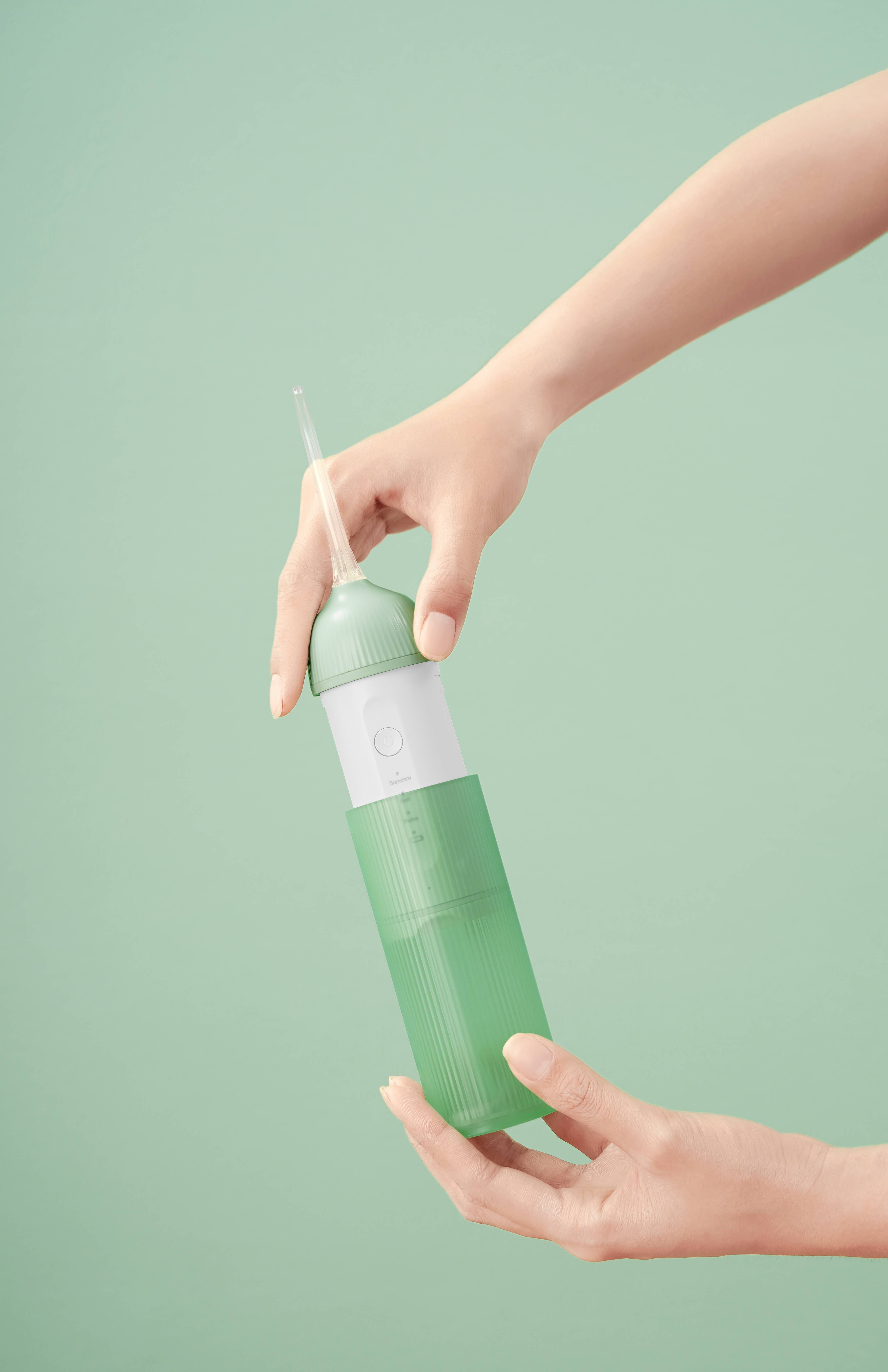 Mani che tengono un idropulsore verde. Forma cilindrica. Parte centrale bianca. Serbatoio trasparente.