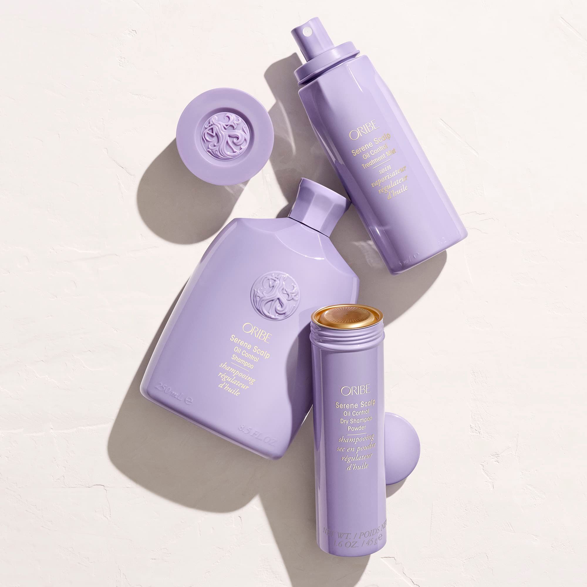 Vari prodotti Oribe. Flaconi e contenitori viola chiaro. Su sfondo chiaro. In alto: flacone spray.