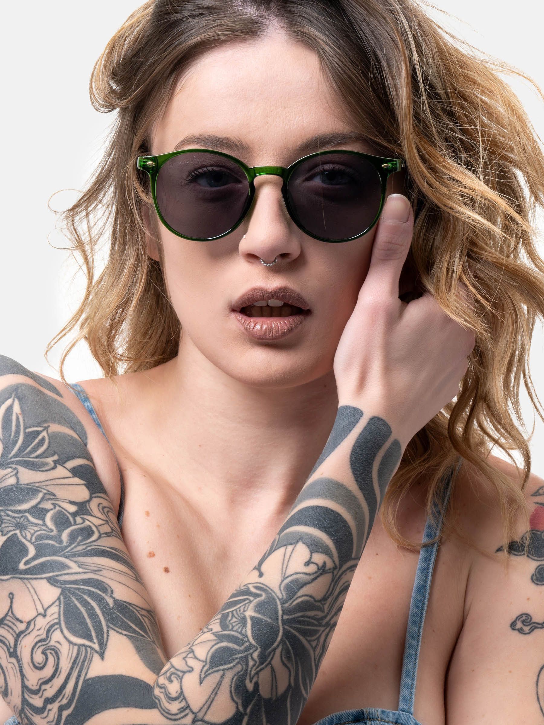 Donna con occhiali da sole verdi. Lenti rotonde, montatura verde. La donna ha tatuaggi sulle braccia.