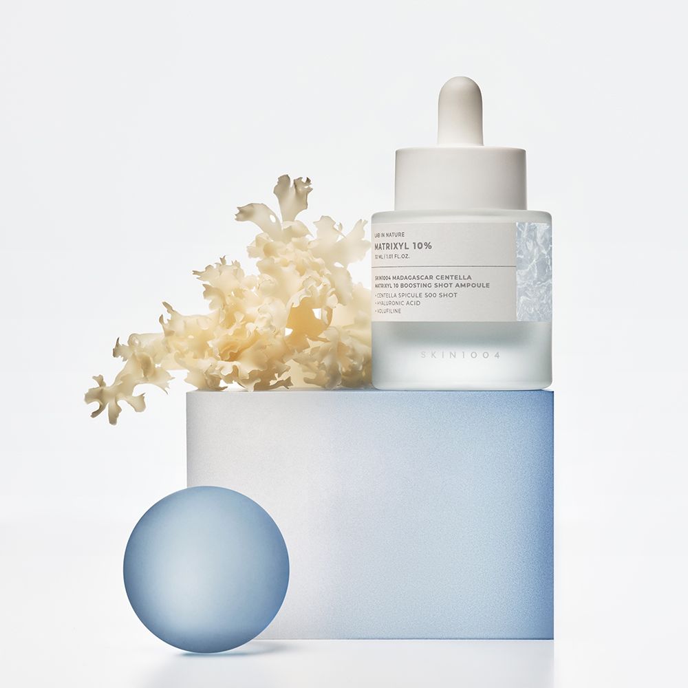 Flacone bianco con pipetta. Scritta: MATRIXYL 10%. Su piedistallo blu e bianco. Decorazione: fiori bianchi e sfera blu.
