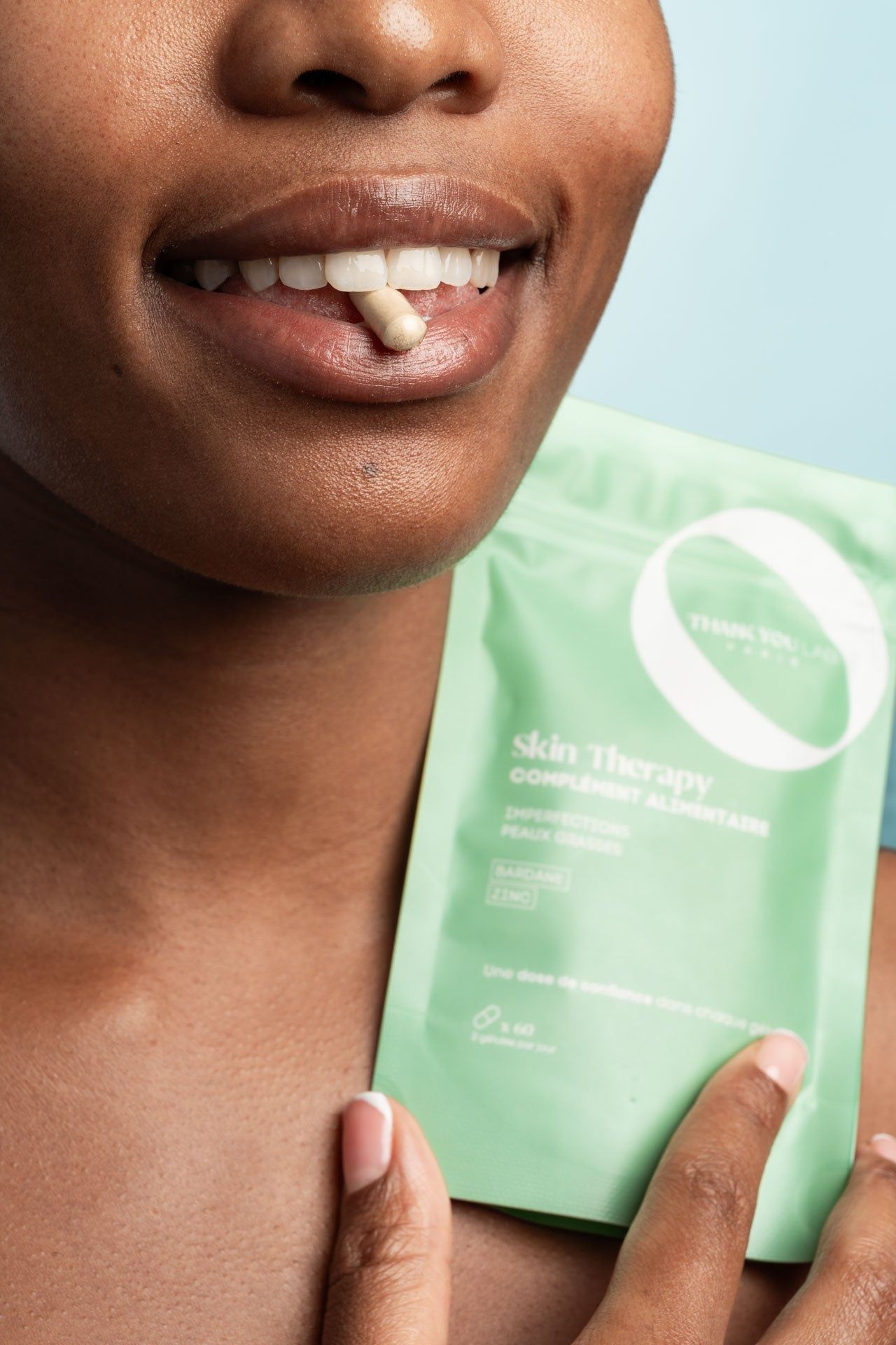 Persona con una capsula in bocca e una confezione verde. Testo: Skin Therapy, Complément Alimentaire.