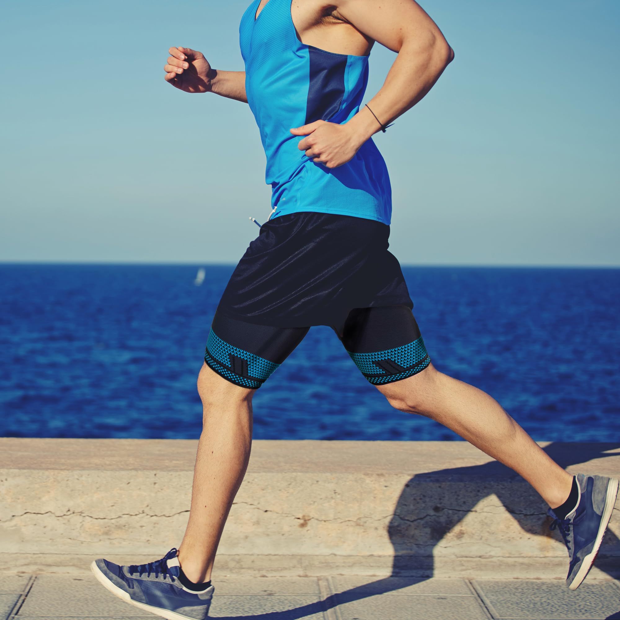Uomo che corre in riva al mare. Pantaloncini neri e bendaggi per cosce blu-neri. Cielo blu e mare sullo sfondo.